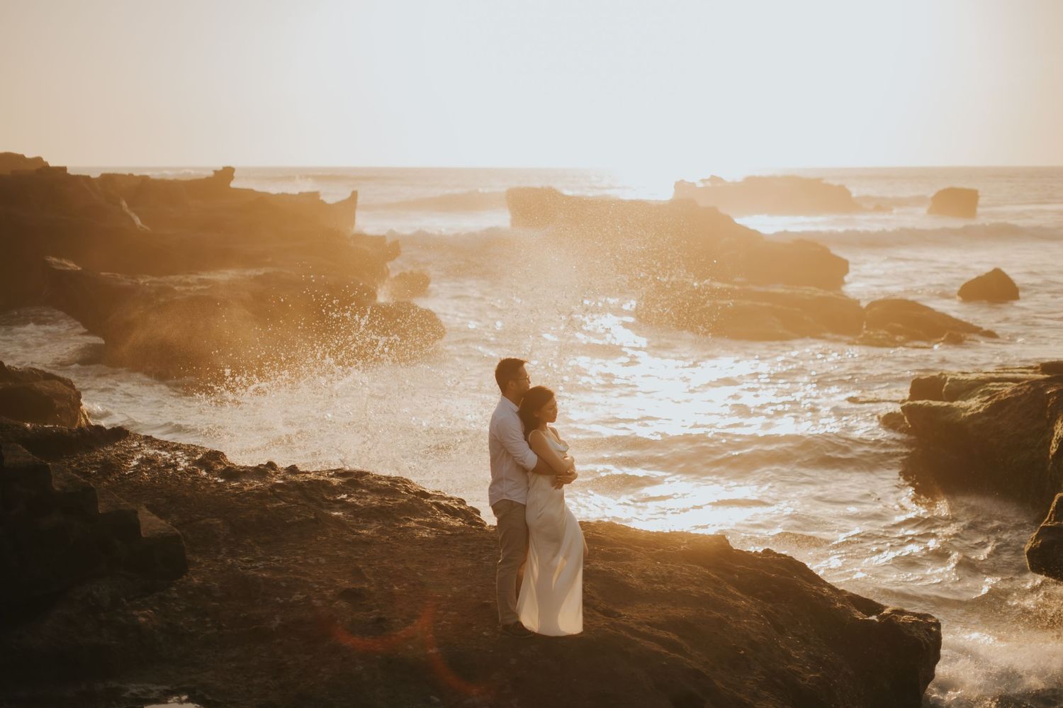 Couple embracing on rocky ocean shore at golden sunset with waves crashing behind them.