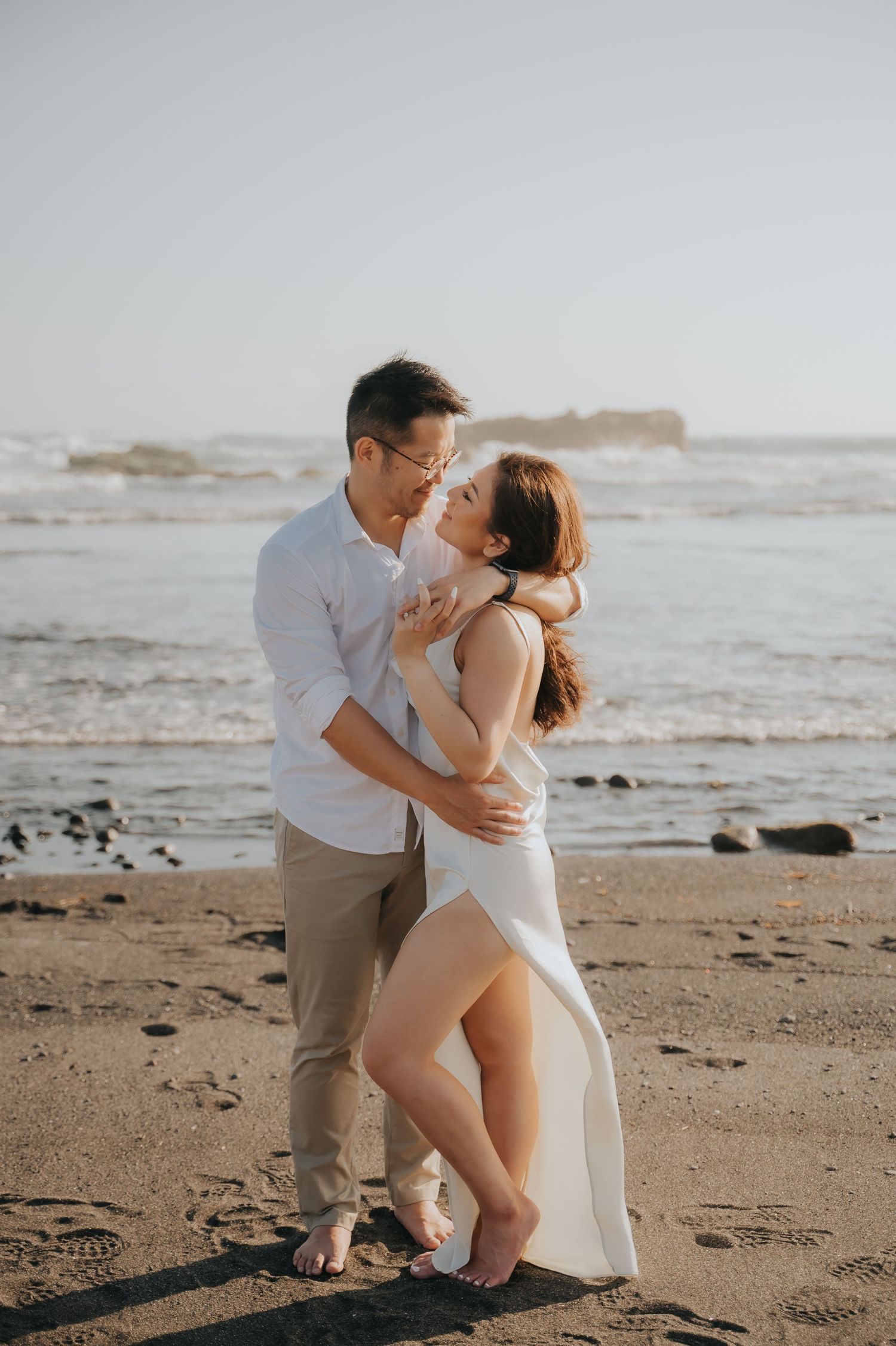 Couple embracing romantically on a sandy beach at sunset, woman in white dress, man in casual attire, ocean waves behind them.