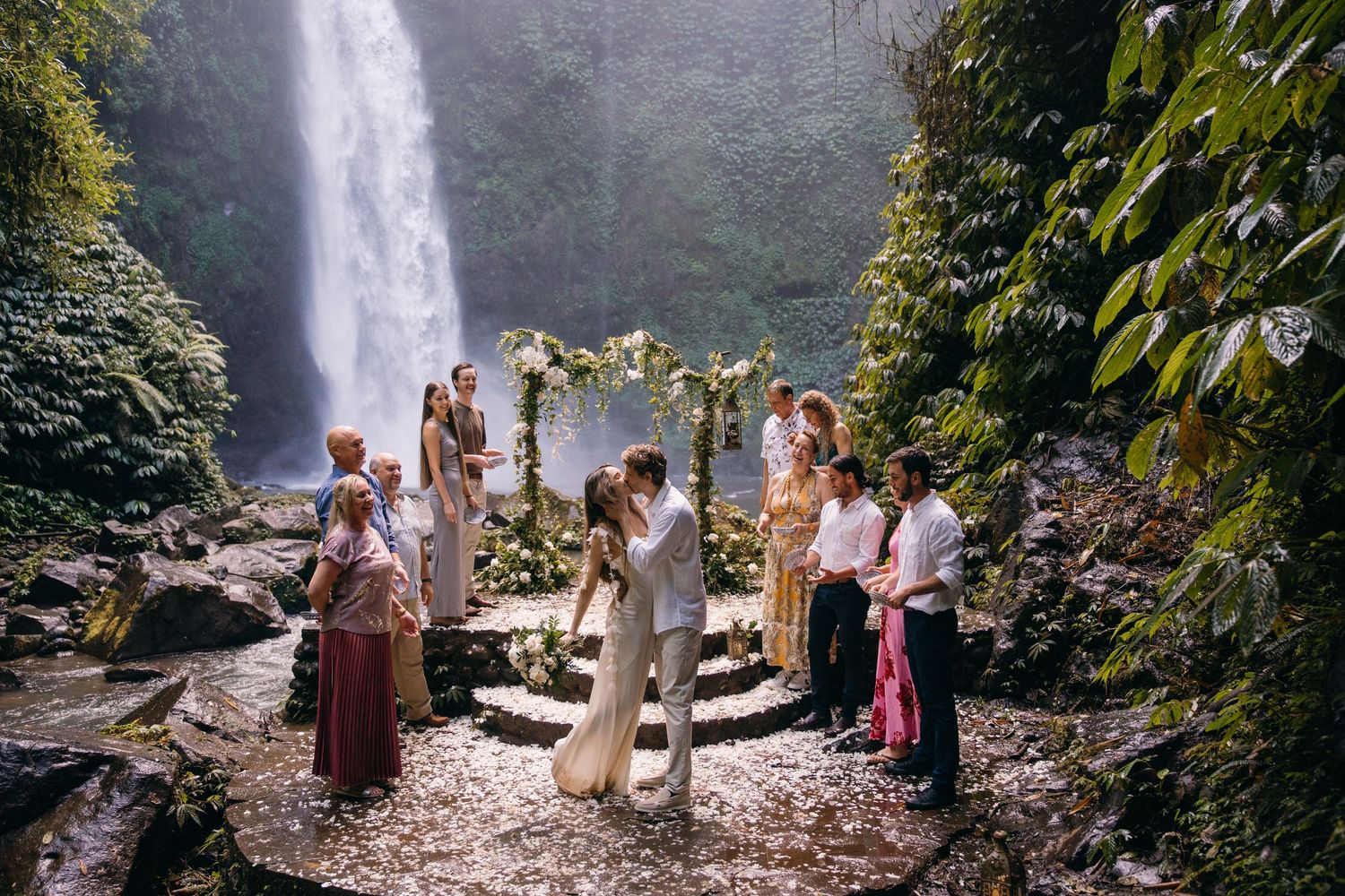 A group of people admiring a scenic waterfall surrounded by lush greenery and rocks in a tropical setting.