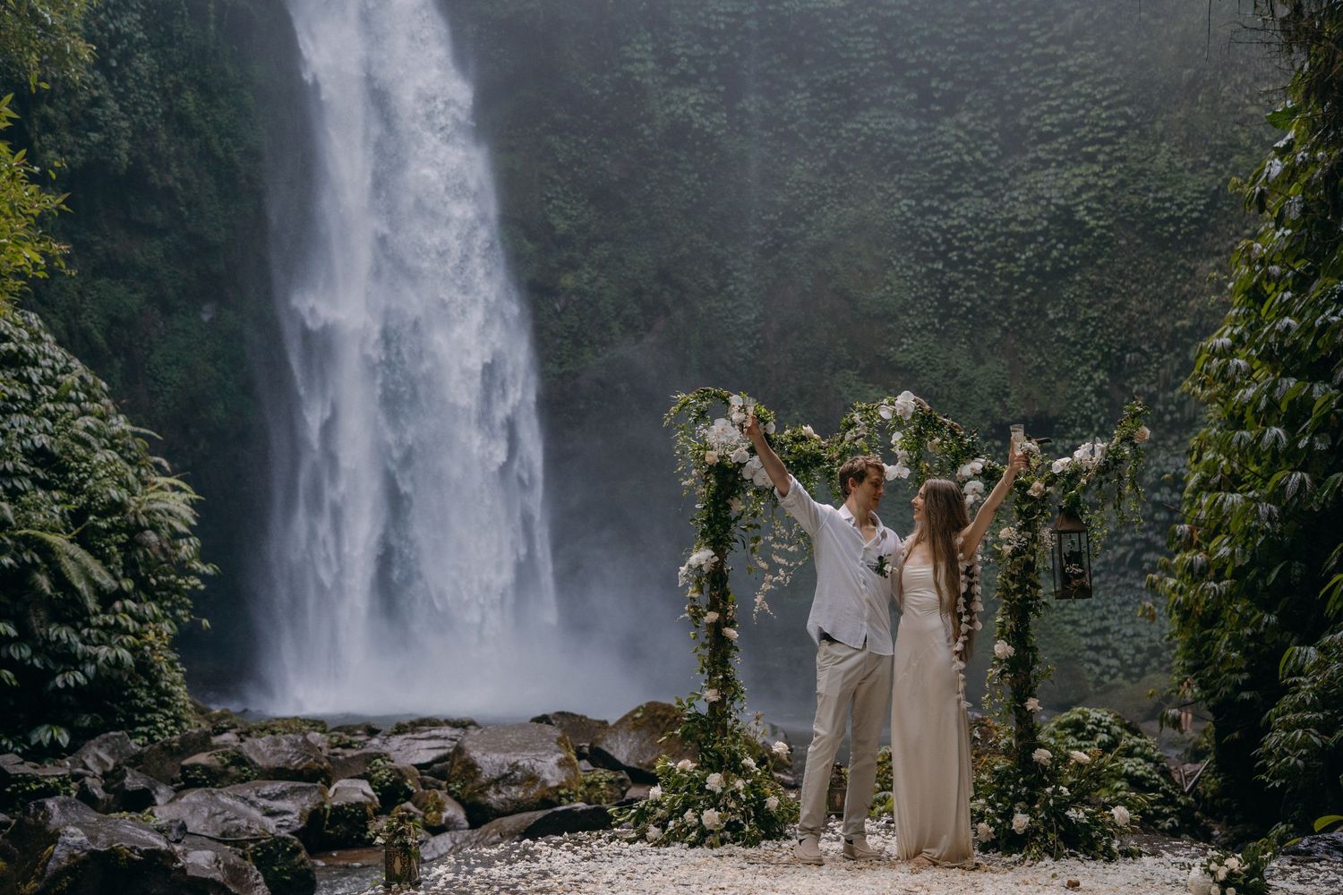 A couple celebrates under a floral arch in front of a majestic tropical waterfall in a lush rainforest setting.