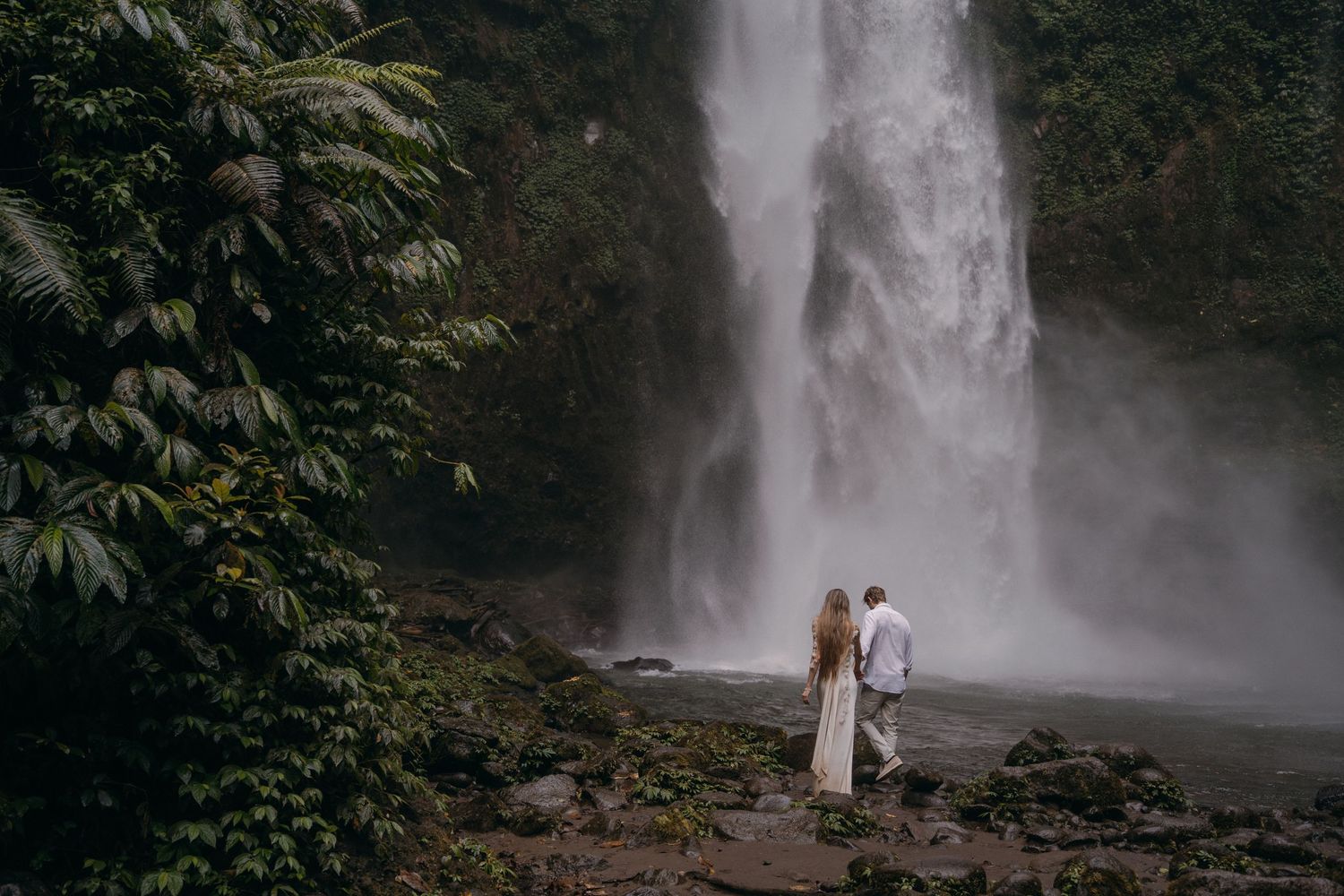 A couple stands in front of a majestic waterfall surrounded by lush tropical foliage in a romantic setting.