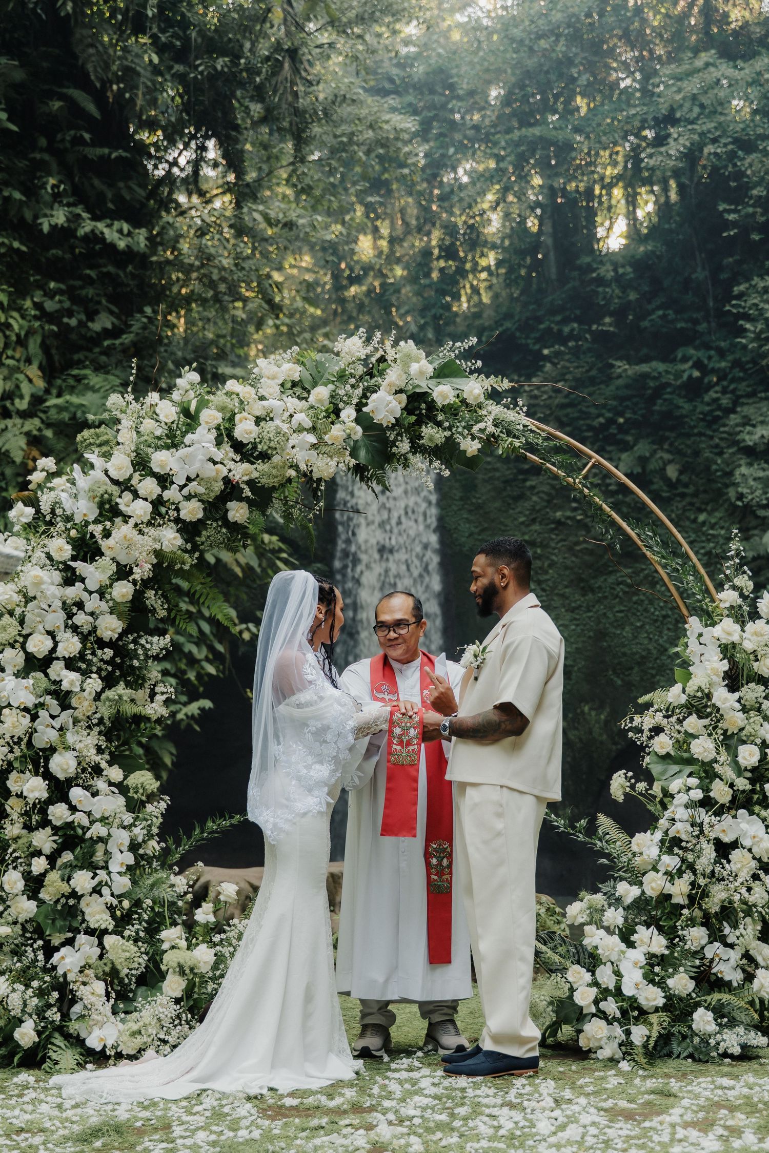 Officiant leading wedding ceremony under white floral arch with stunning waterfall in Bali jungle.