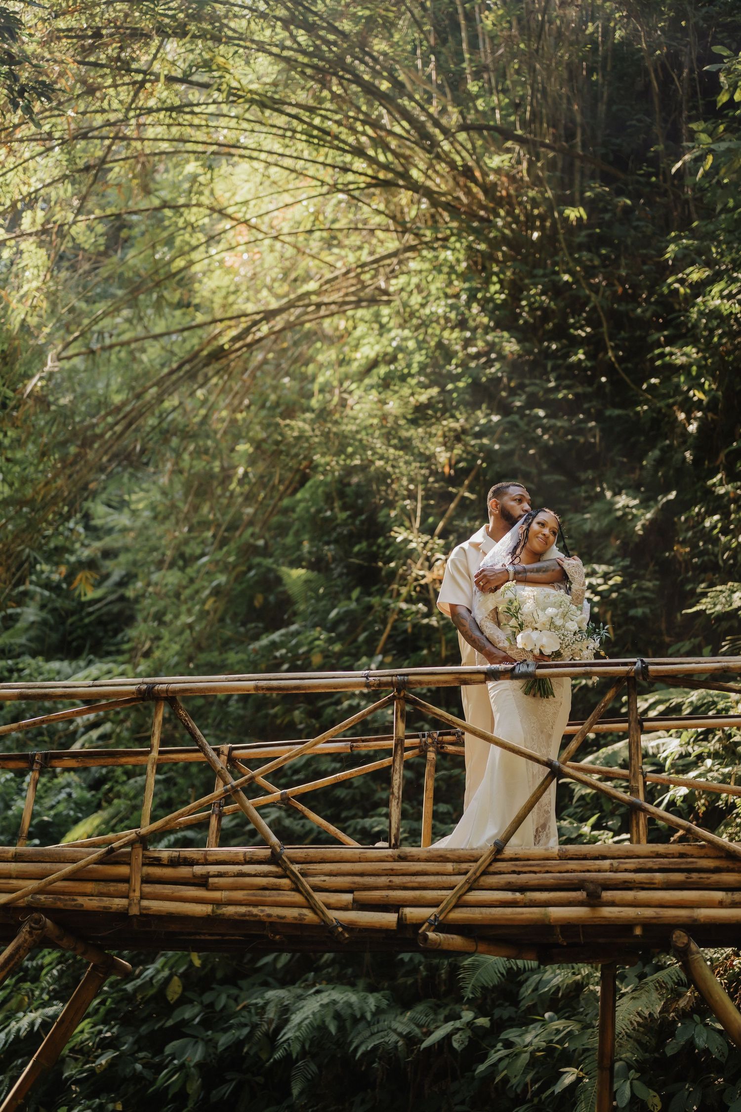 Wedding couple poses romantically on a bamboo bridge surrounded by a sunlit tropical rainforest setting.