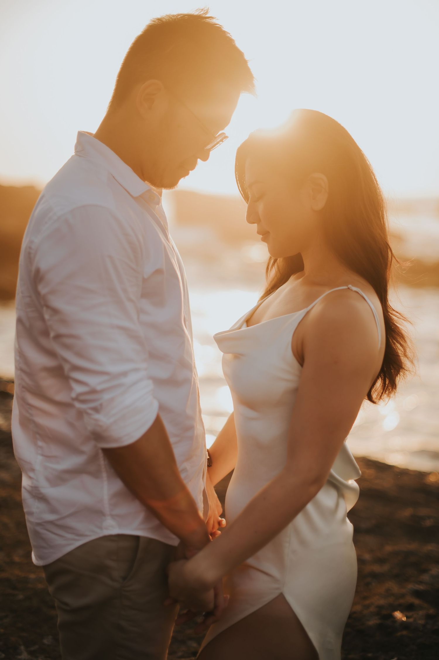 Couple holding hands at golden sunset by the water, woman in white dress and man in white shirt sharing intimate moment.