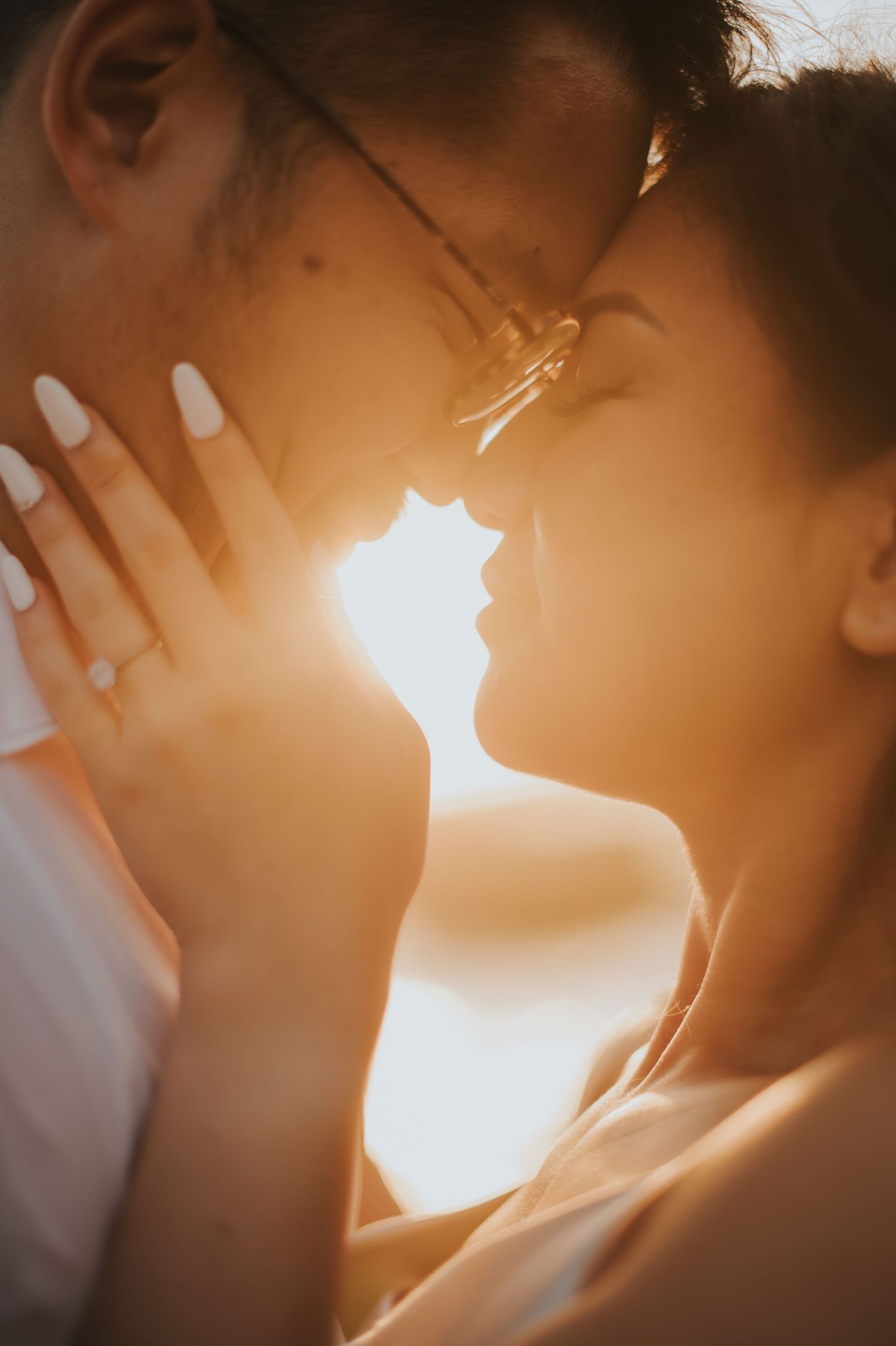 Couple sharing a romantic moment at golden sunset, woman with white nails and glasses leaning in close to partner.