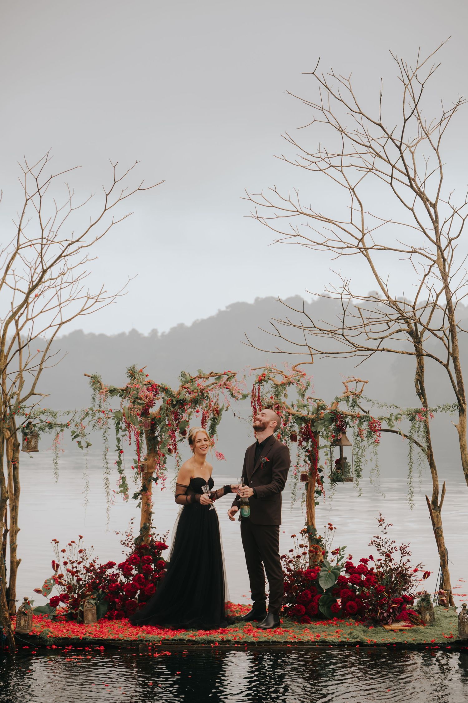 Waterfront wedding ceremony with bare trees and crimson floral decorations creating a dramatic atmosphere.