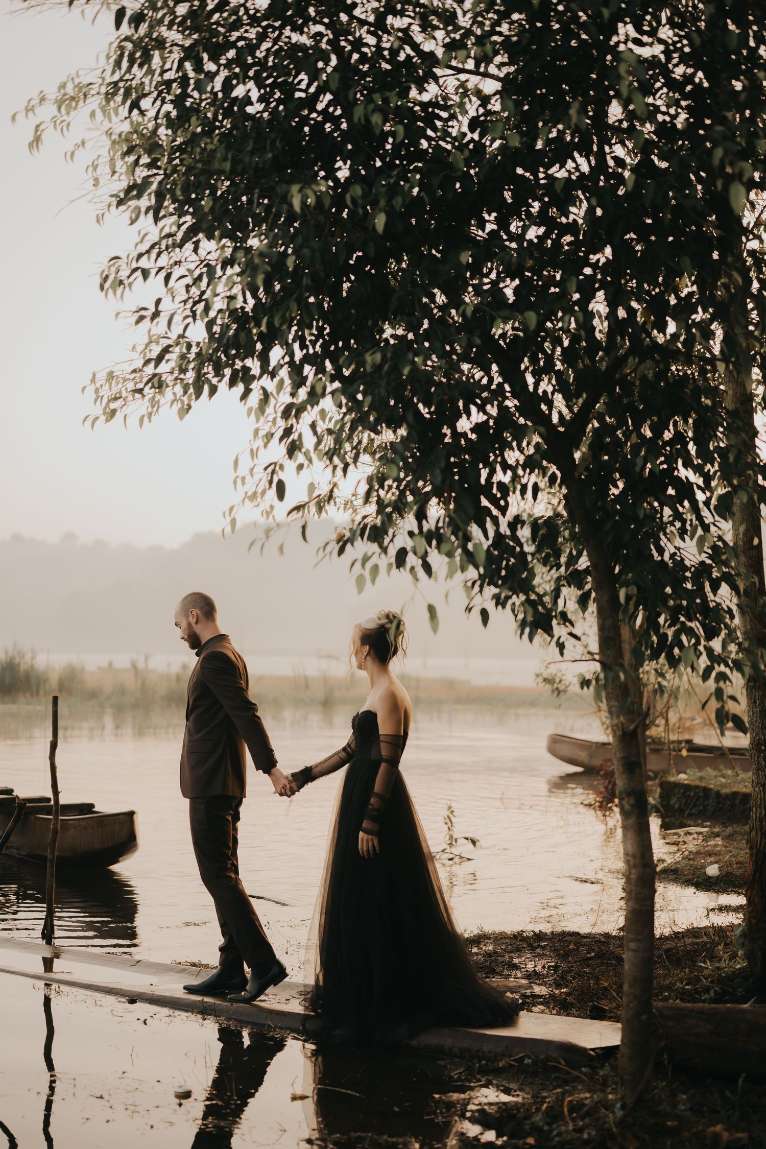 A romantic couple in formal attire stands together on a misty lakeside dock surrounded by trees at sunset.