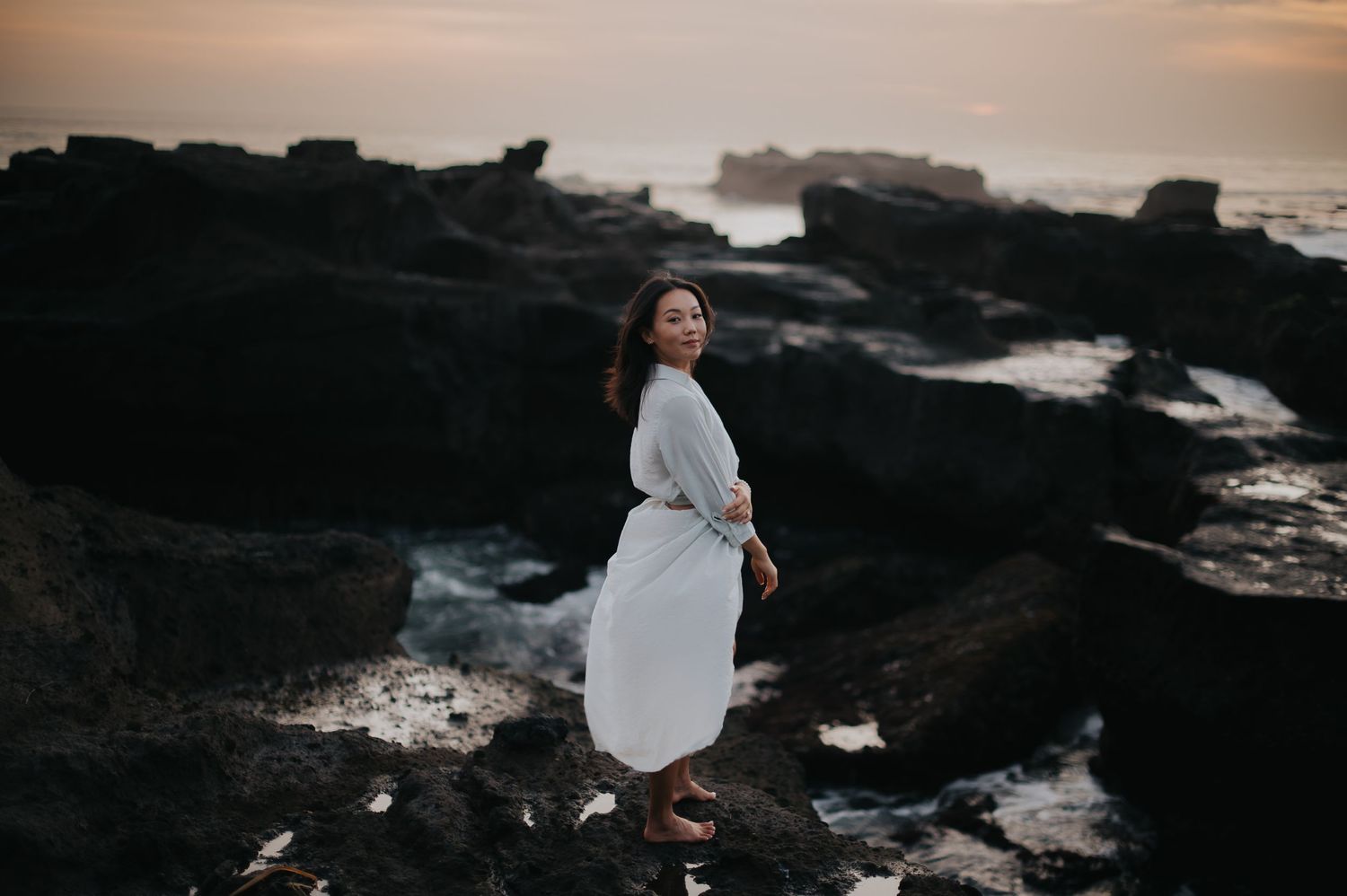 Person in white dress stands on rocky coastal shoreline during golden sunset with waves crashing in background.