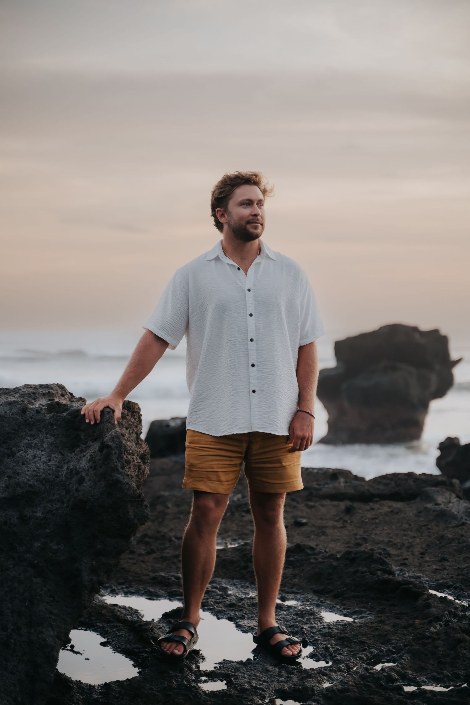Person in white shirt and brown shorts stands on volcanic rocks near ocean during cloudy sunset.