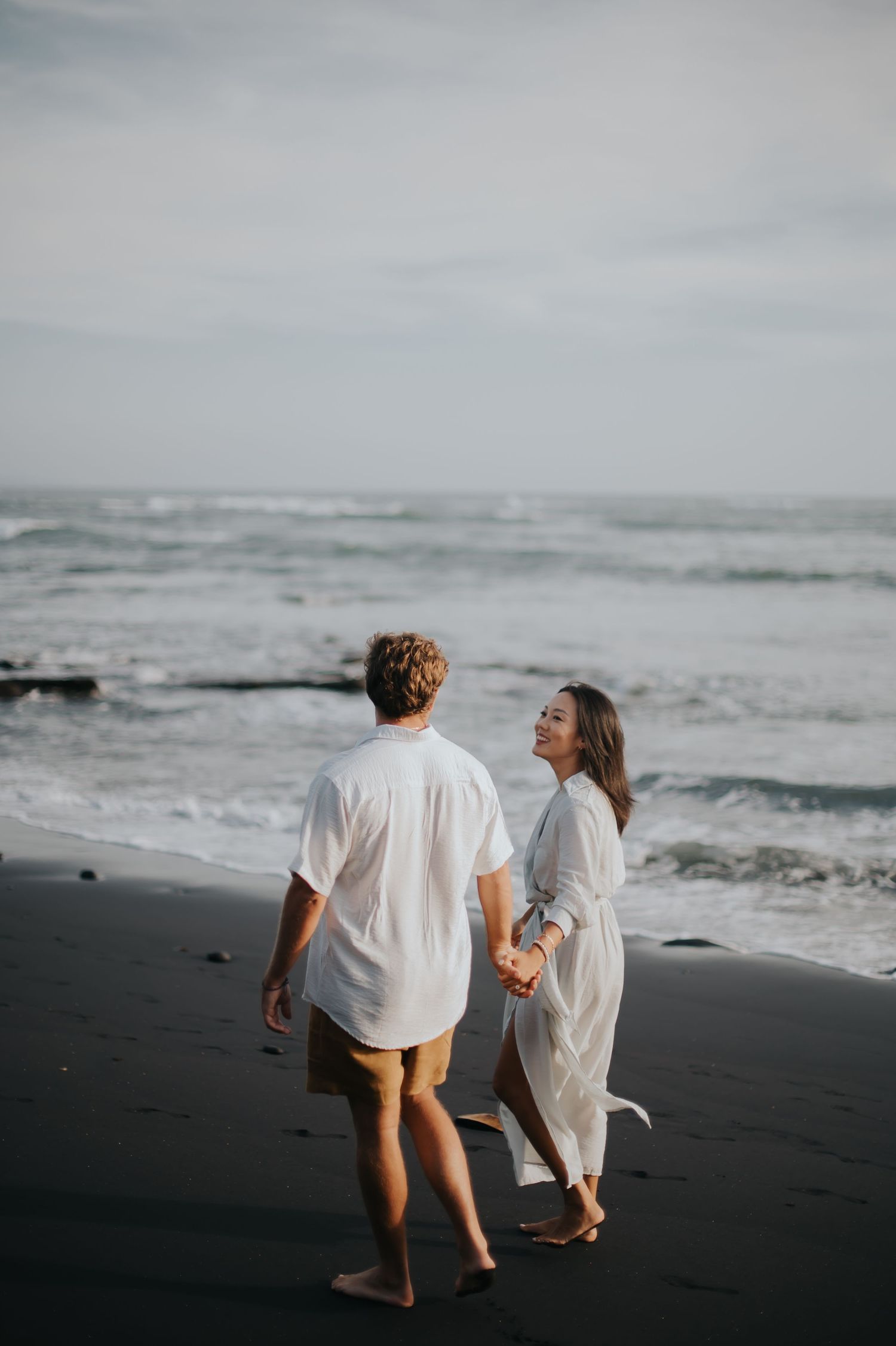Couple walking together on black sand beach holding hands with ocean waves in background.