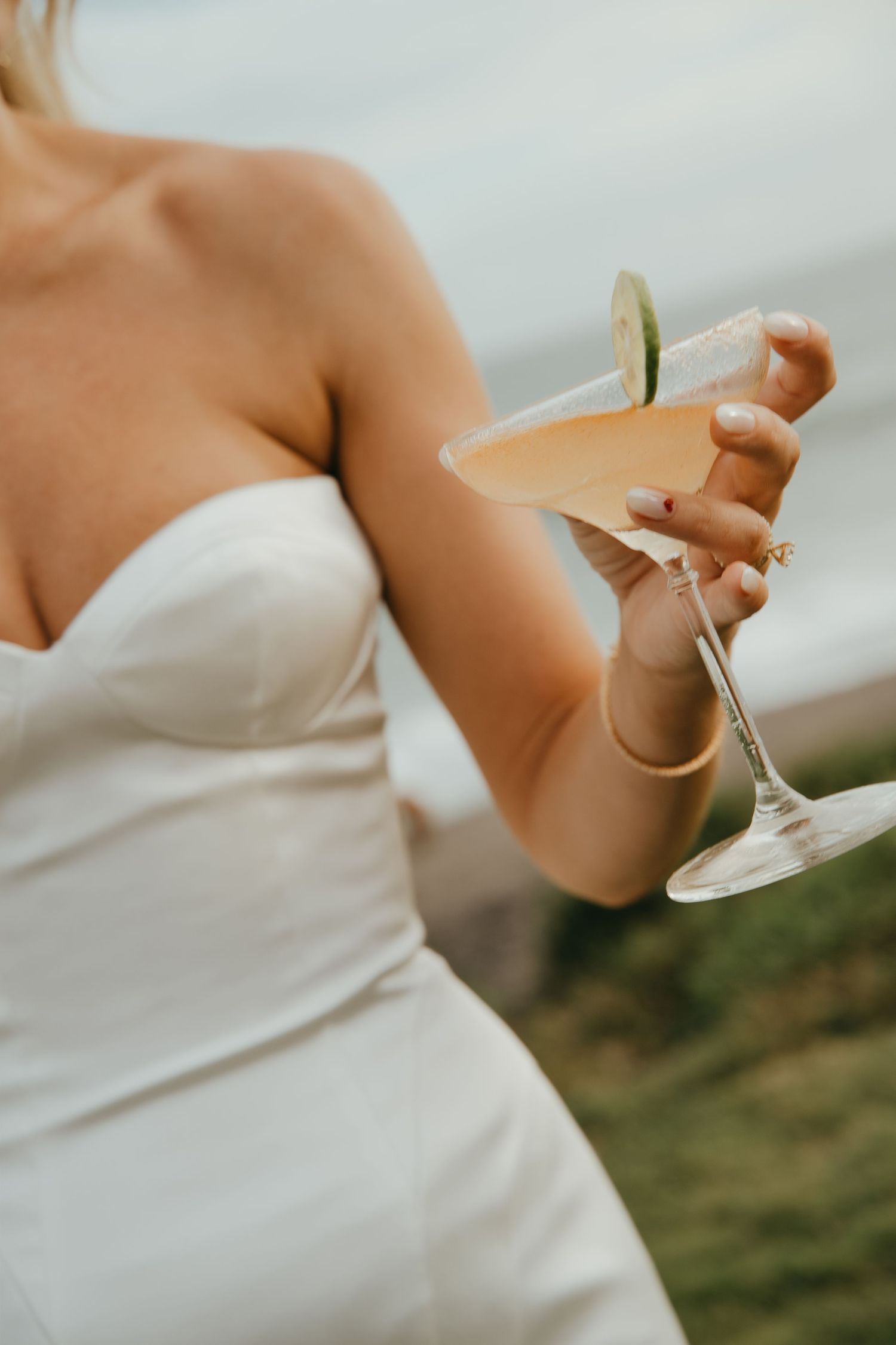 Bride in strapless white gown holding a cocktail garnished with lime at outdoor wedding.