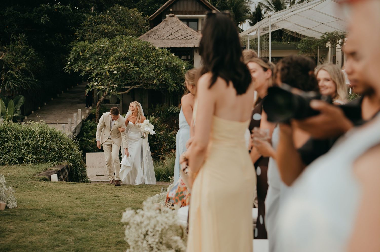 Bride and groom walking down outdoor aisle surrounded by wedding guests and bridesmaids in yellow.