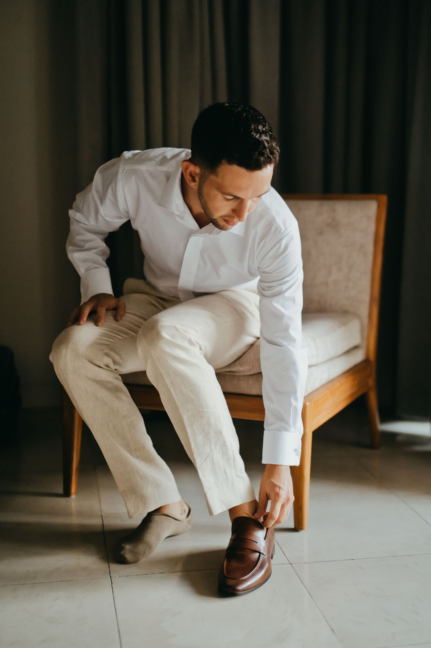 Groom in white shirt and cream trousers putting on brown loafers while seated on elegant chair.