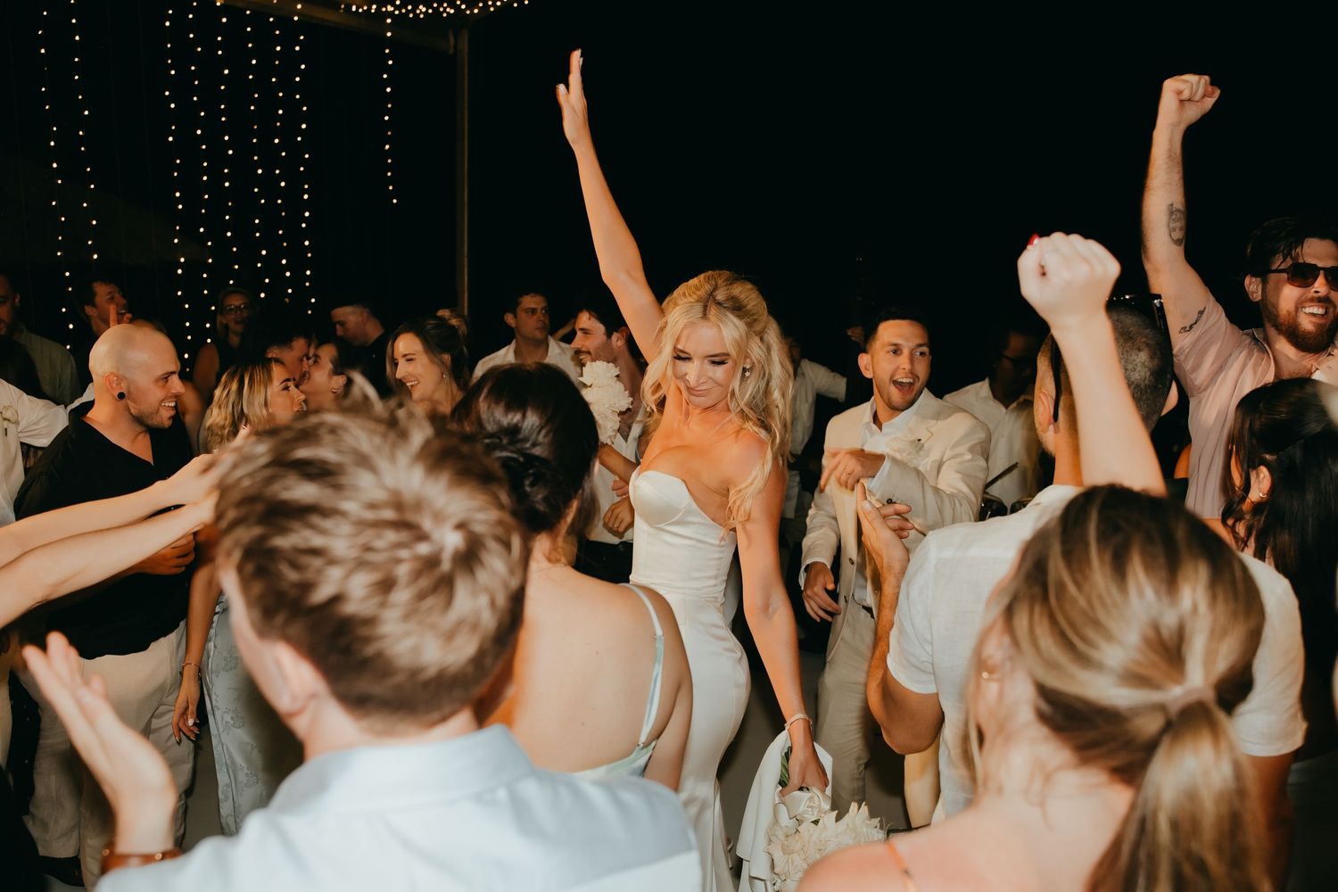 Bride in fitted white gown dances joyfully with arms raised surrounded by cheering wedding guests at reception.