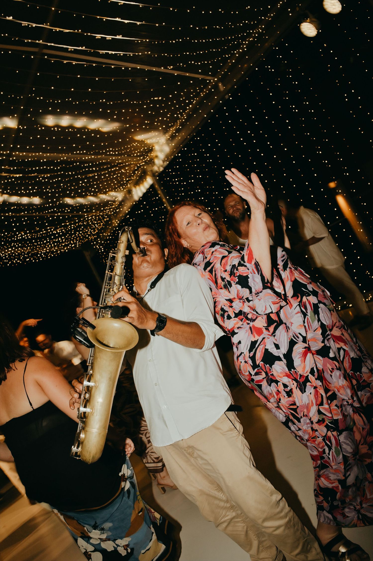 Saxophonist performing on the dance floor at a lively wedding reception under fairy lights.