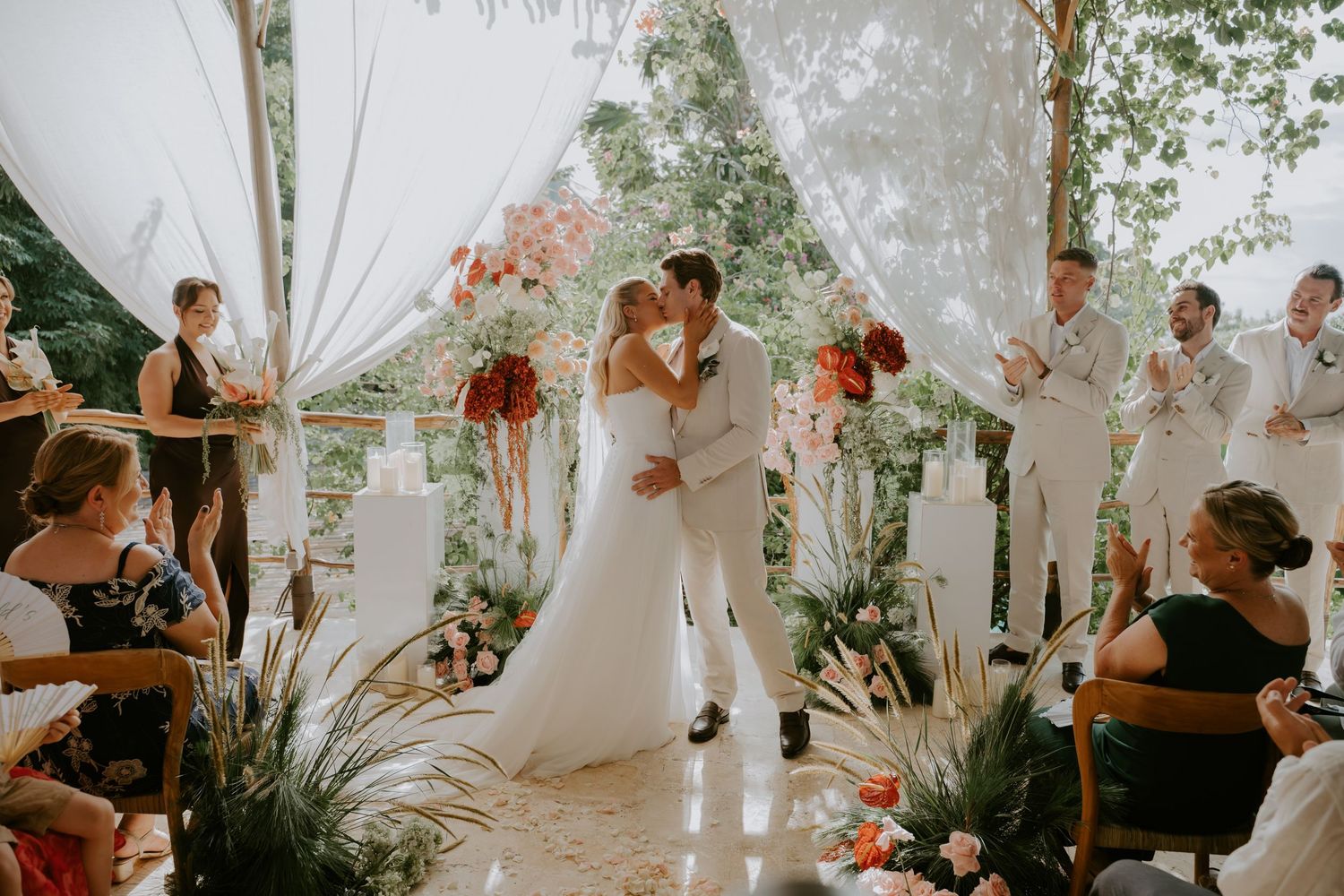Bride and groom share first kiss at outdoor bohemian wedding ceremony with floral arch and white draping.