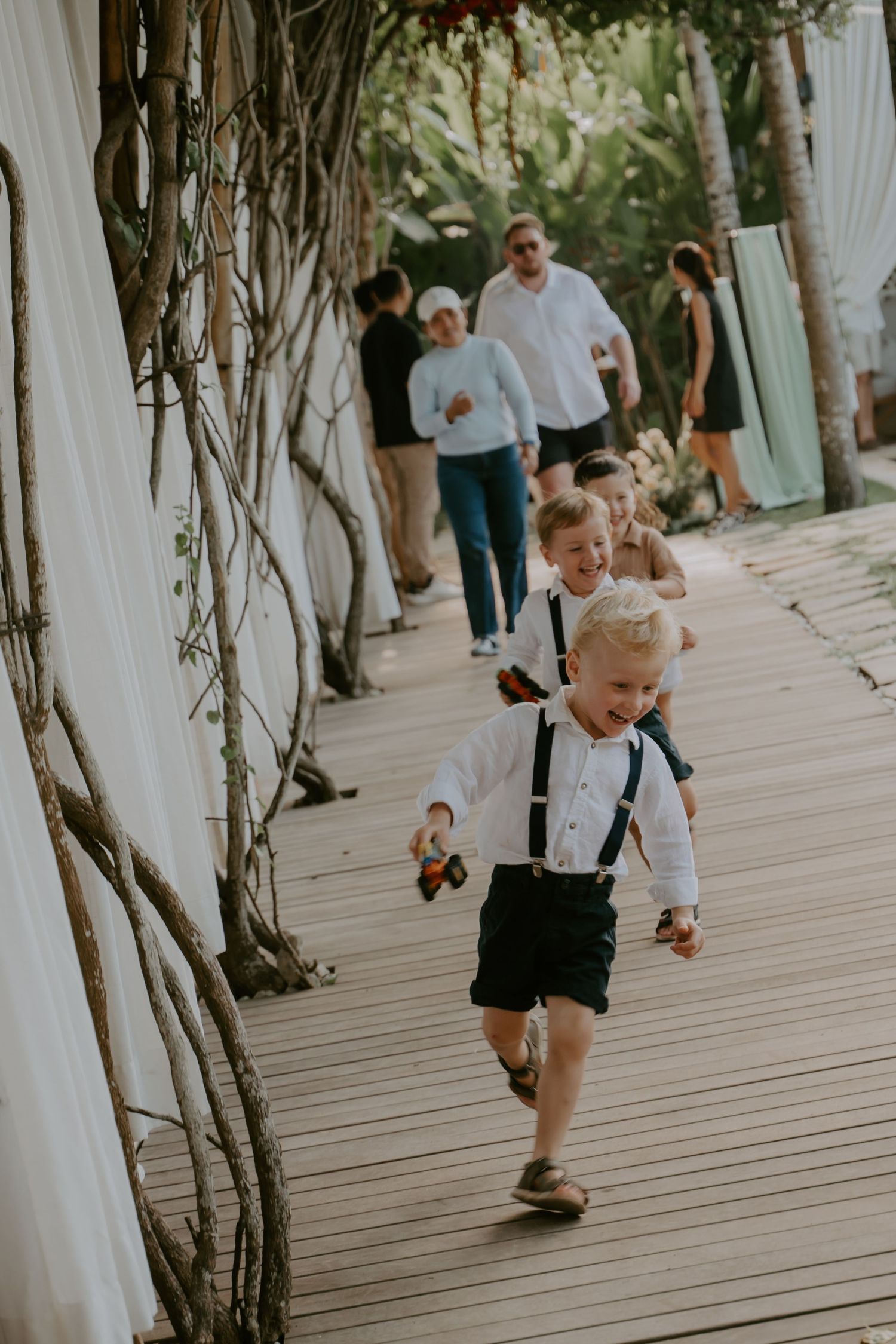 Young ring bearer in white shirt and suspenders runs down wooden aisle at outdoor wedding ceremony.