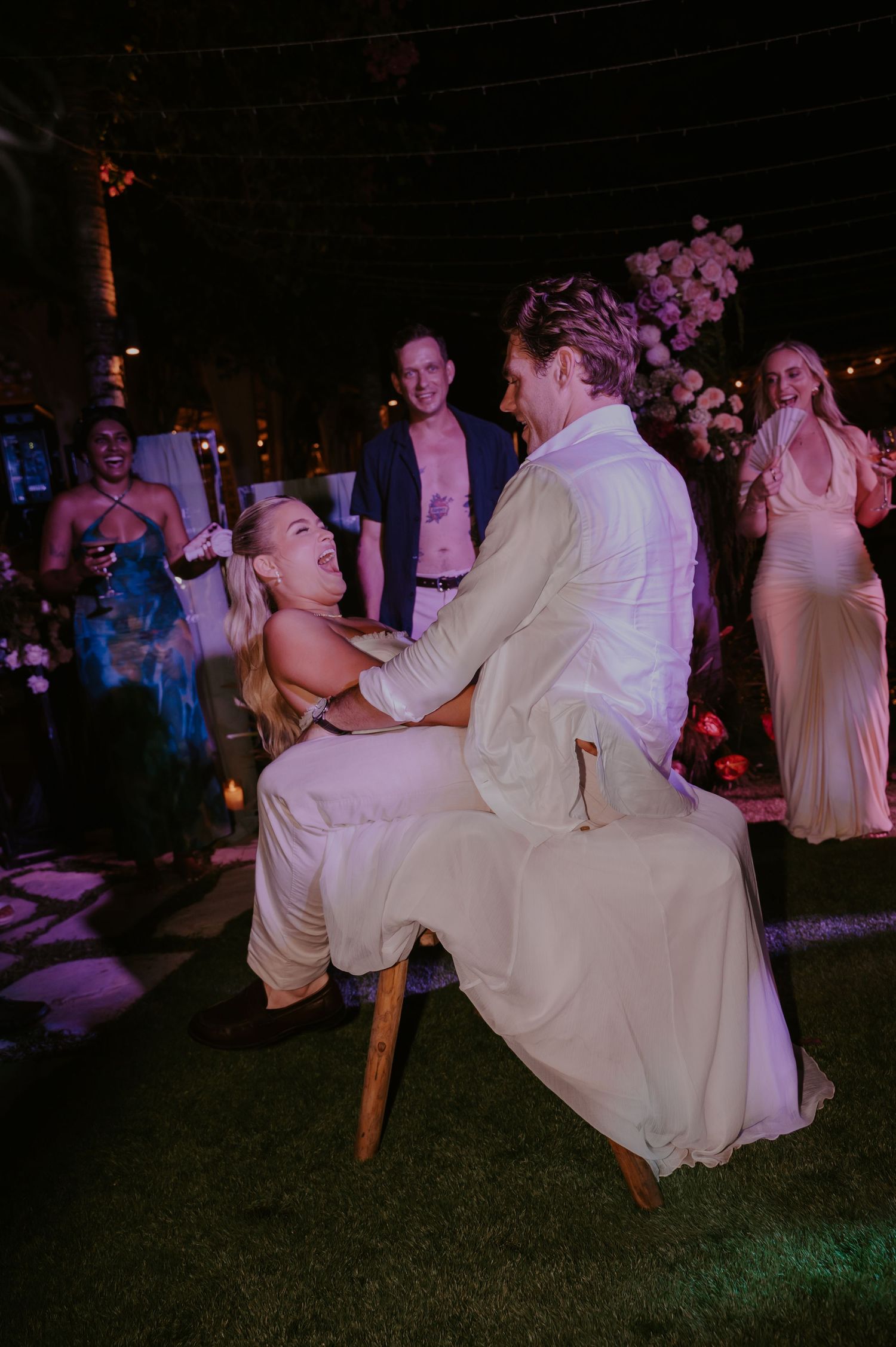 Bride kneeling before groom during fun garter removal tradition at tropical outdoor wedding reception.