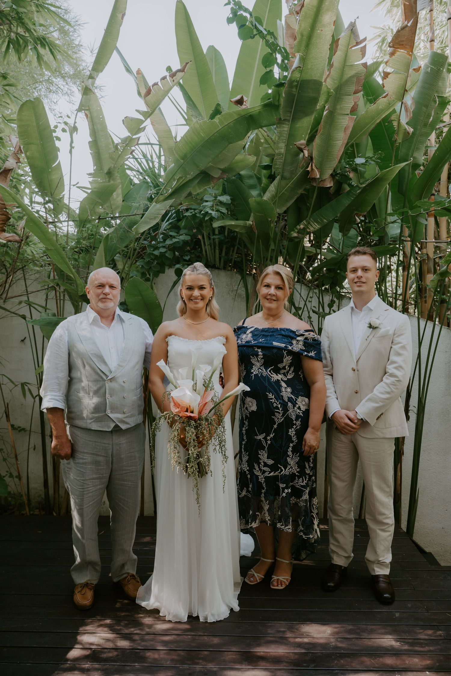 Bride and groom posing with parents in tropical garden setting, bride holding colorful tropical bouquet.