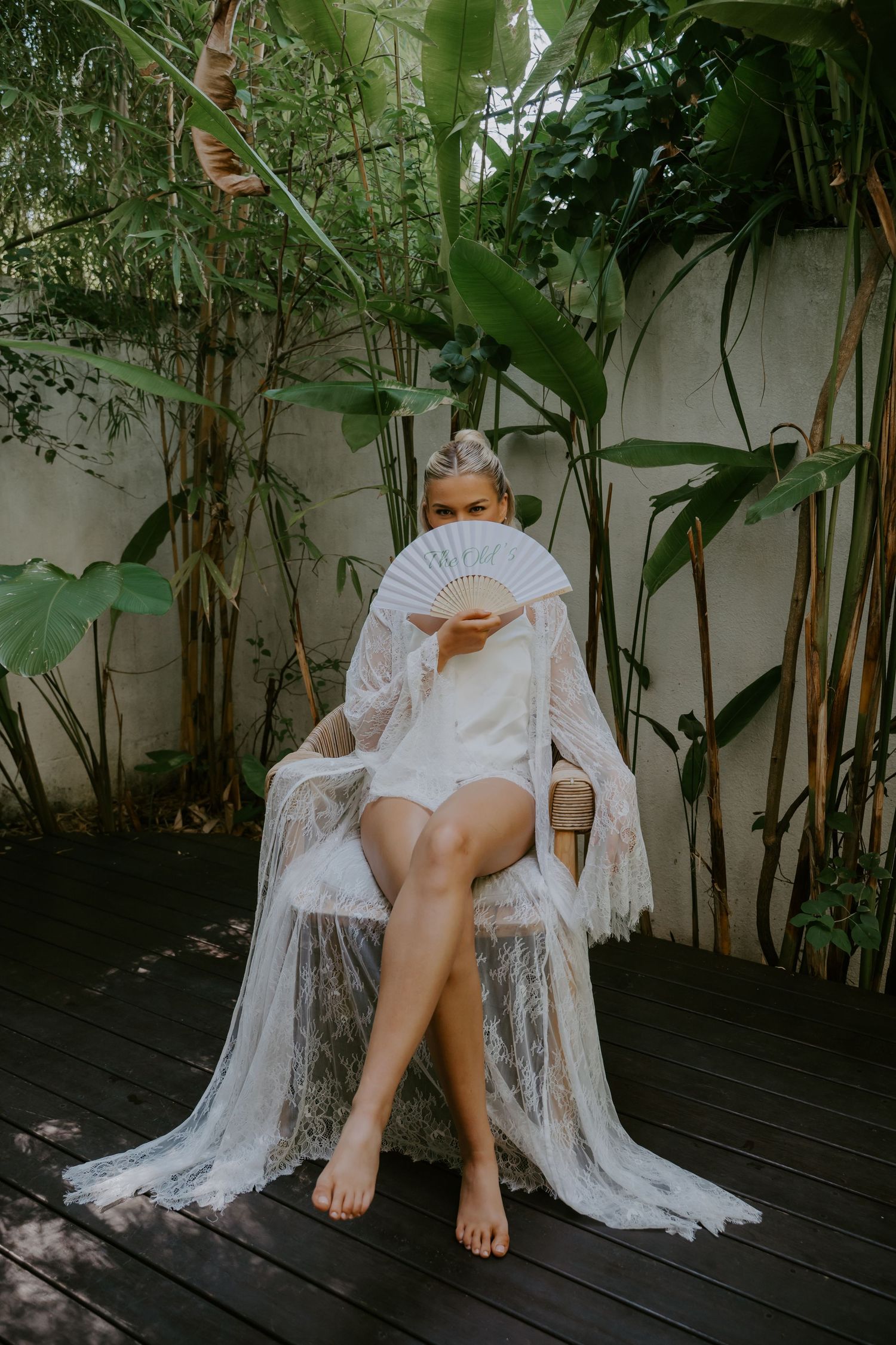 Bride in white lace robe sits in a rattan chair holding a decorative fan surrounded by tropical plants.