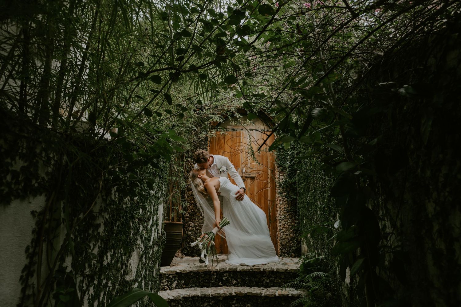 Bride and groom kissing in a romantic ivy-covered archway with wooden door.