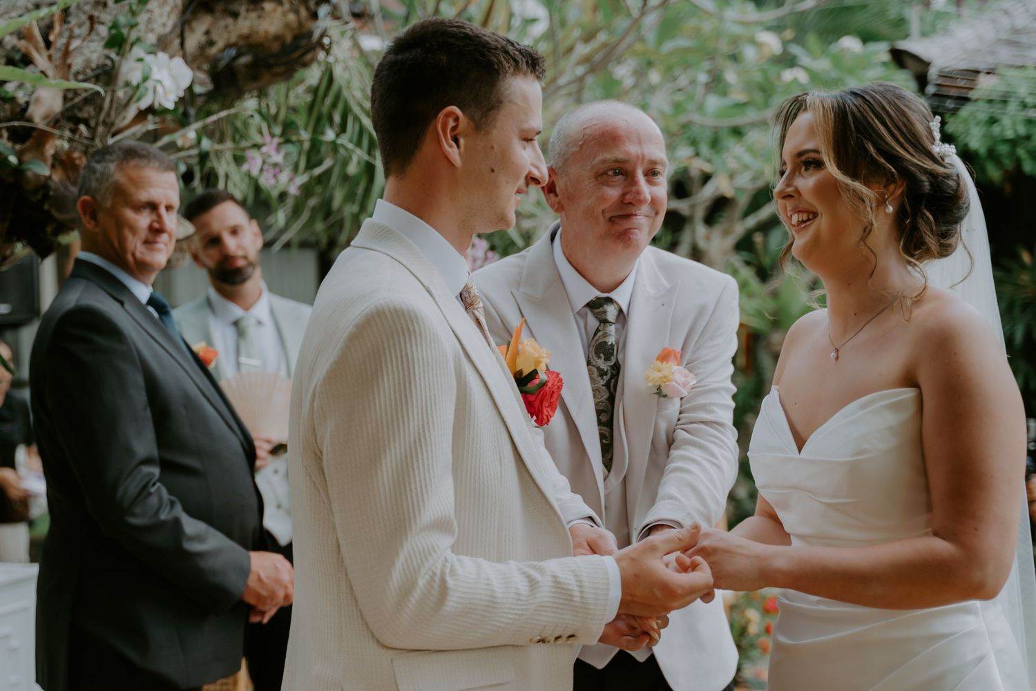 Groom in white suit and bride in strapless gown exchanging vows at outdoor Bali wedding ceremony.