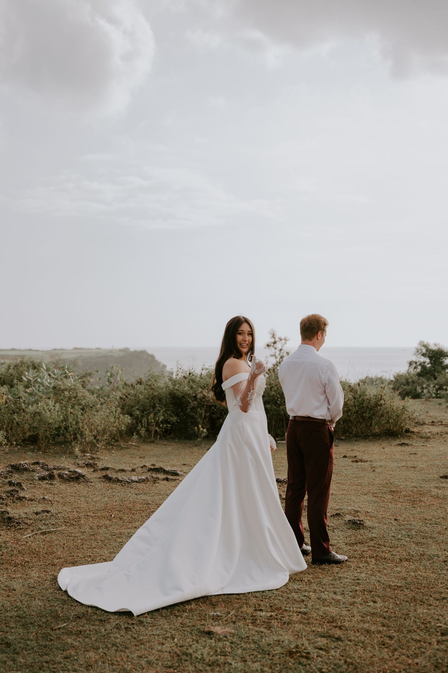 Bride in white off-shoulder gown with train looks back while groom stands facing ocean at outdoor wedding.