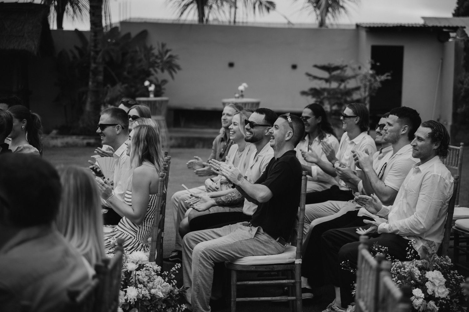 Wedding guests laughing and clapping during outdoor ceremony in black and white photo.