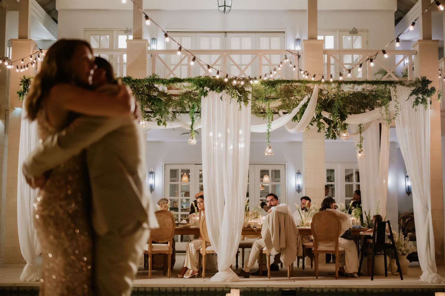 Couple sharing romantic first dance at elegant outdoor wedding reception with string lights and greenery.