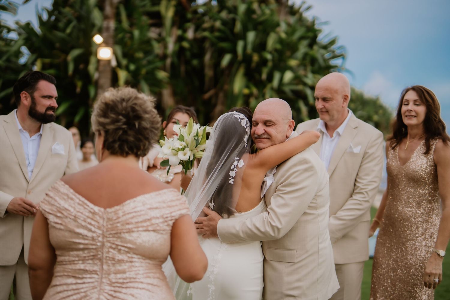 Emotional groom embraces guest as bride in veil looks on during outdoor wedding ceremony.