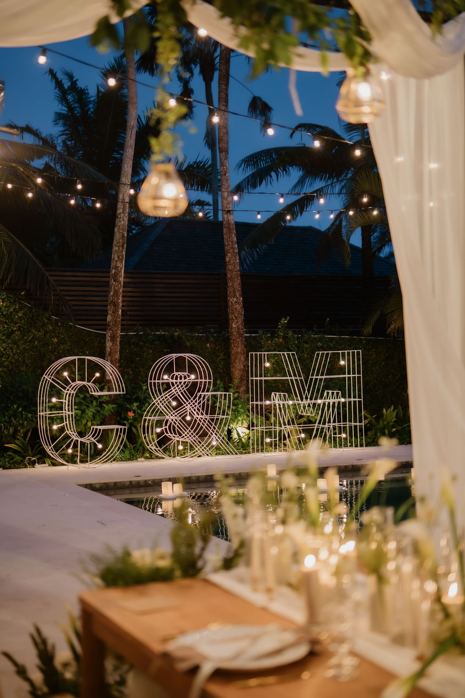 Wedding reception with illuminated C&M marquee letters beside pool with string lights at night.