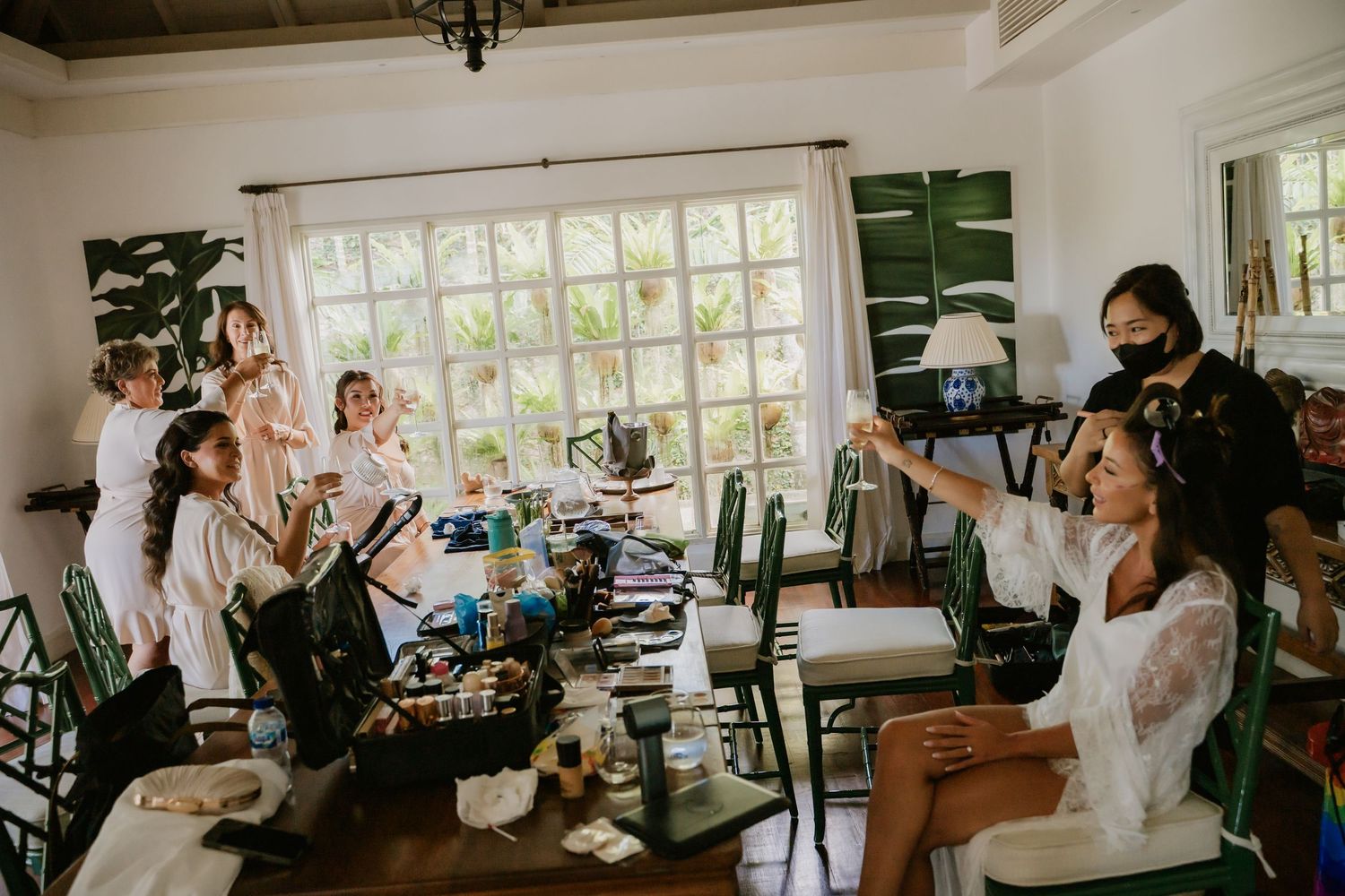 Bridal party getting ready in elegant room with green shutters, bridesmaids in white robes with makeup artist working.