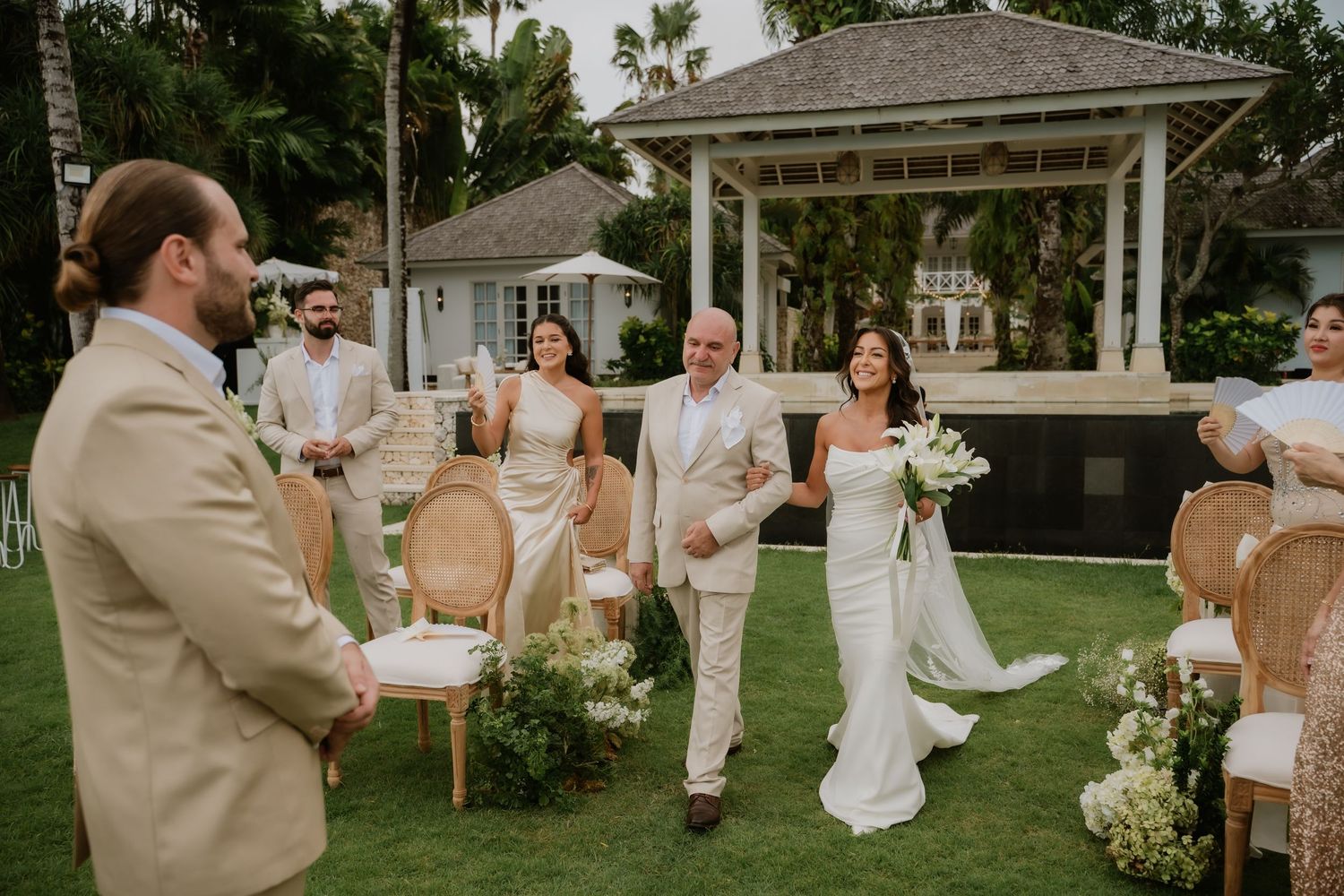 Bride in white gown walks down outdoor aisle with escort at tropical wedding ceremony near gazebo.