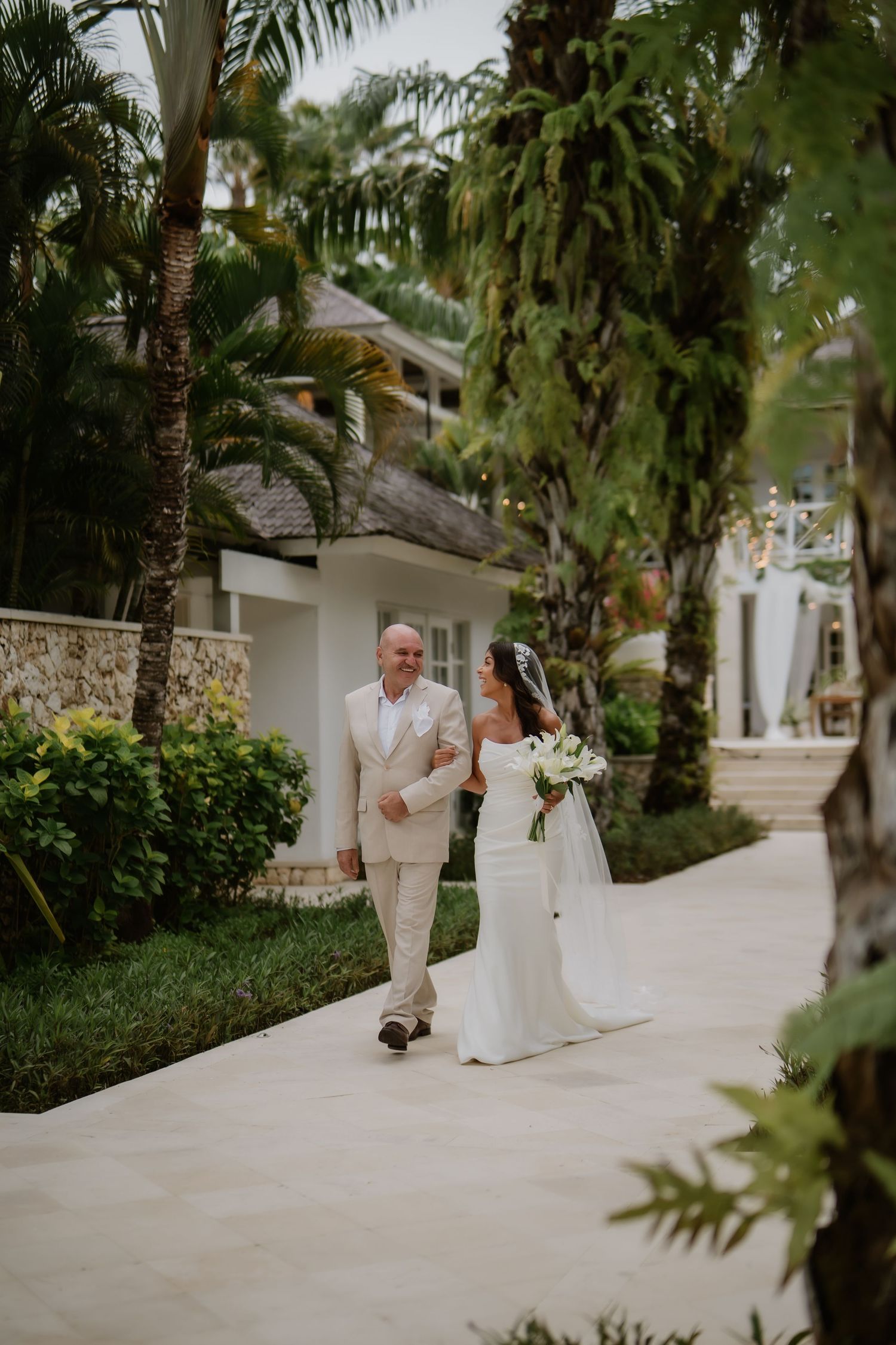 Bride in strapless white gown walking with father along palm-lined pathway at tropical resort venue.