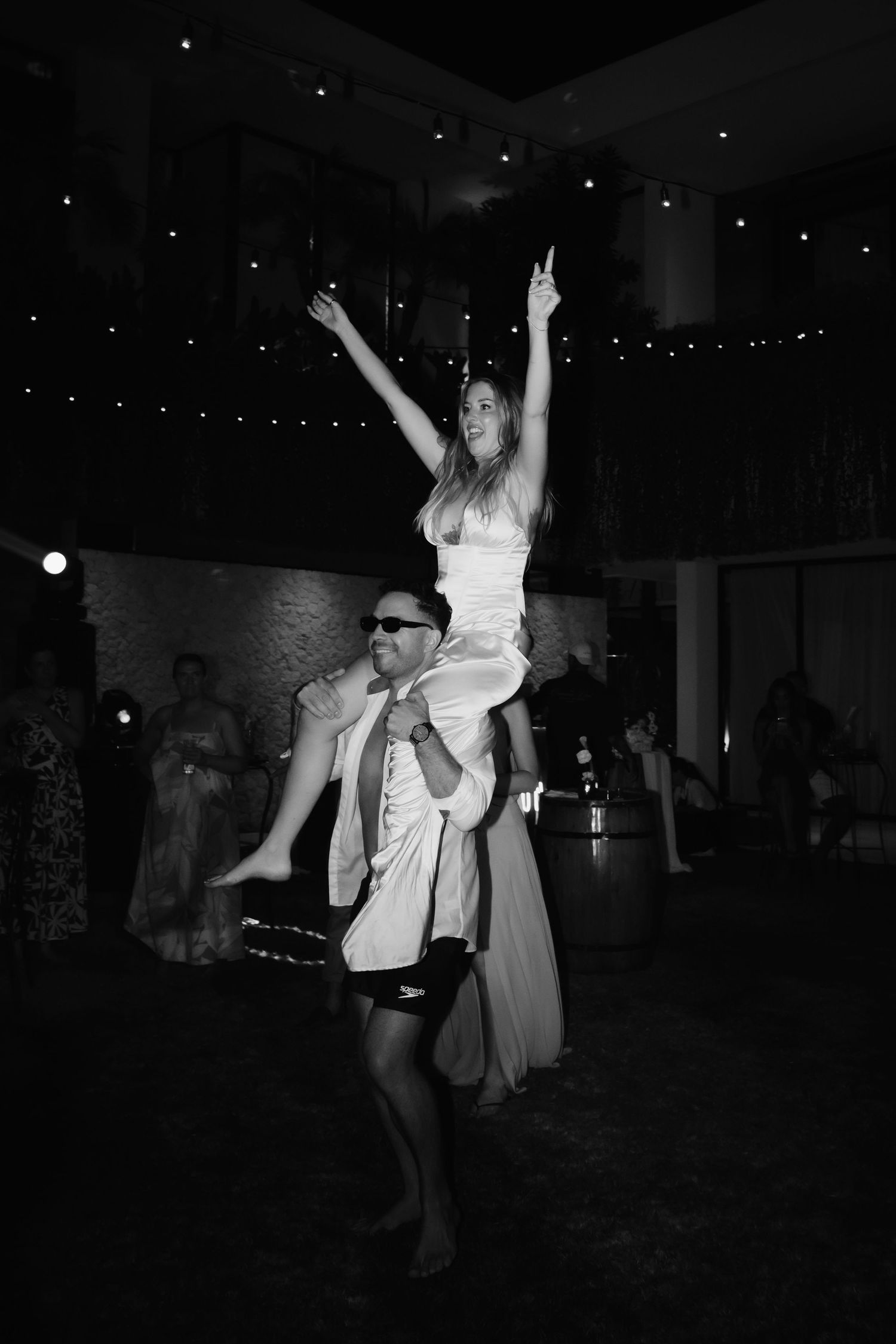 Black and white photo of bride lifted on shoulders dancing joyfully at wedding reception.