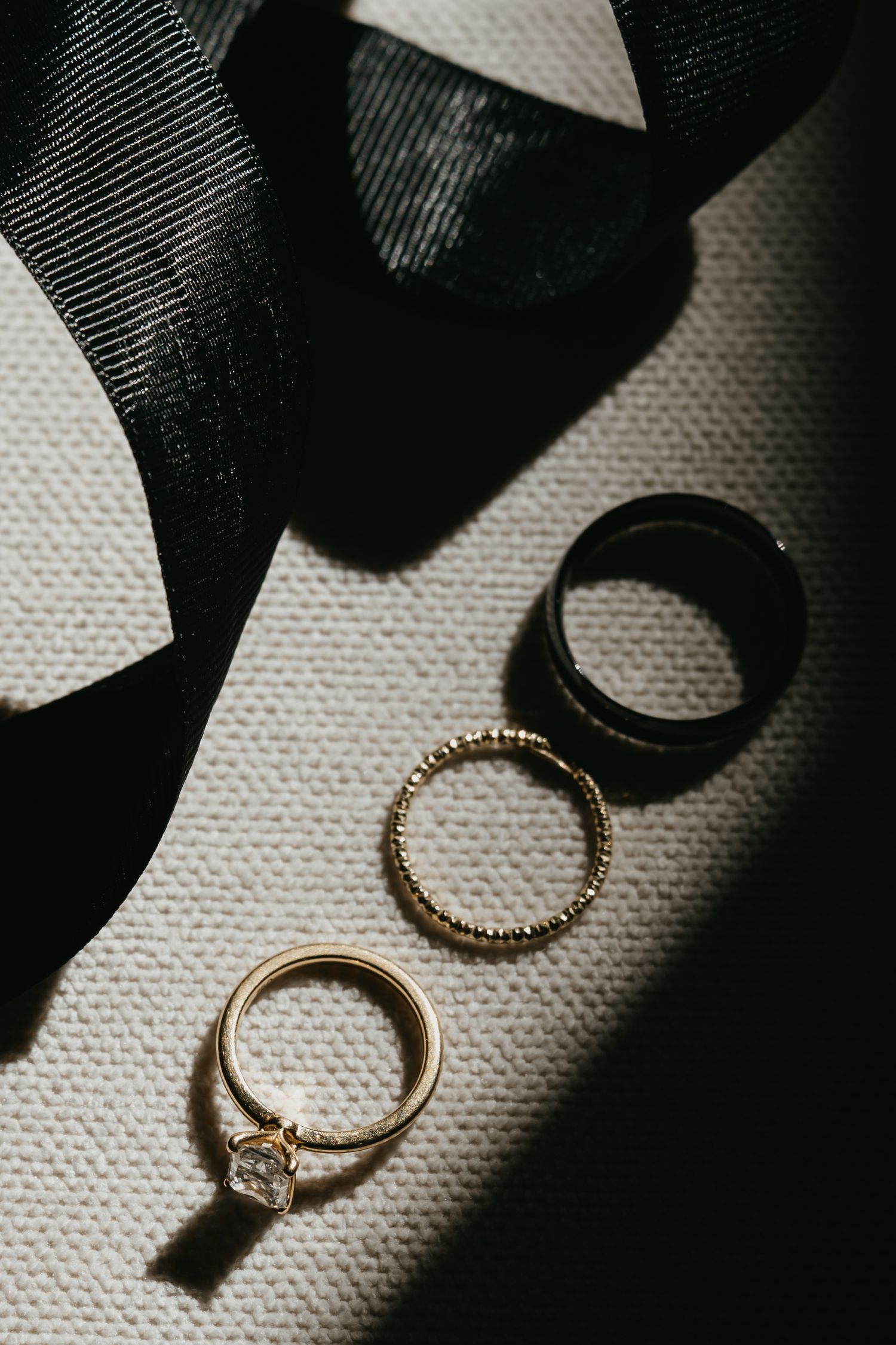 Three wedding rings, including a diamond solitaire, gold band, and black ring, arranged on white linen with black ribbon.