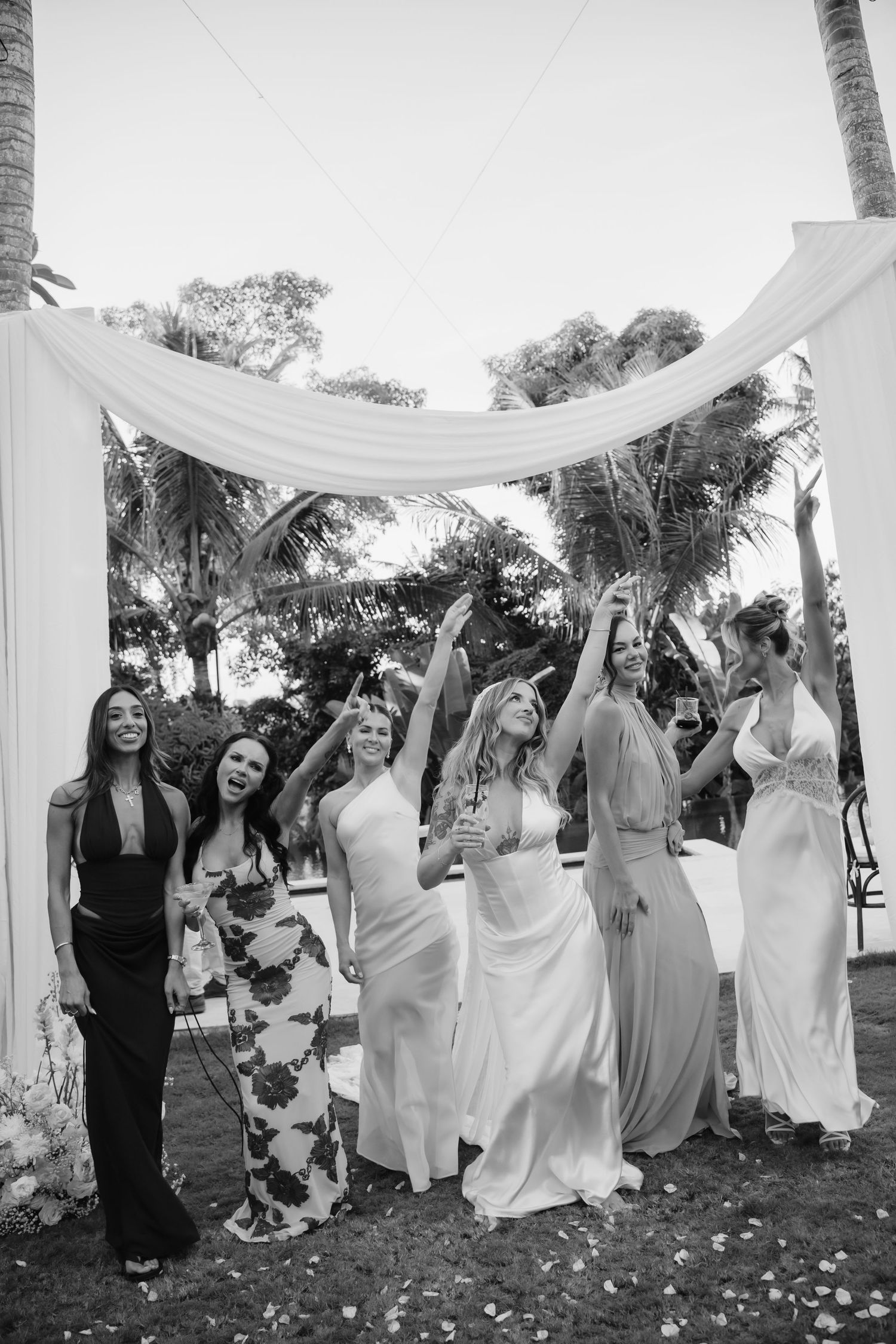 Black and white photo of bride and bridesmaids celebrating under floral arch at outdoor wedding.