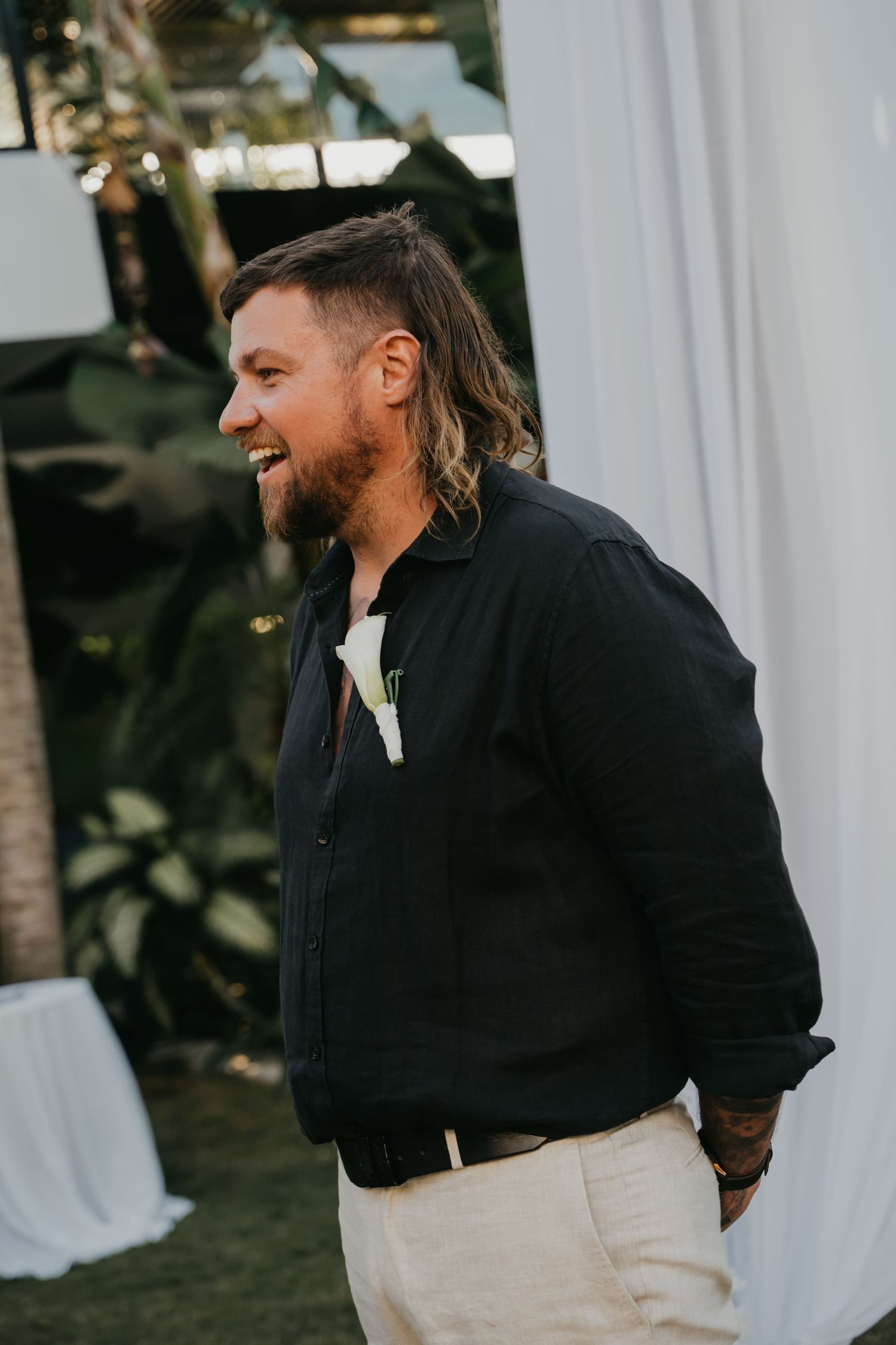 Smiling bearded groom with long hair in black linen shirt and khaki pants at tropical outdoor wedding ceremony.