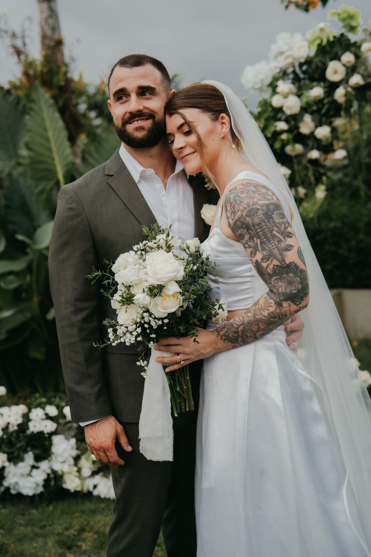 Newlyweds posing together outdoors, bride with tattoo sleeve holding white rose bouquet in garden.