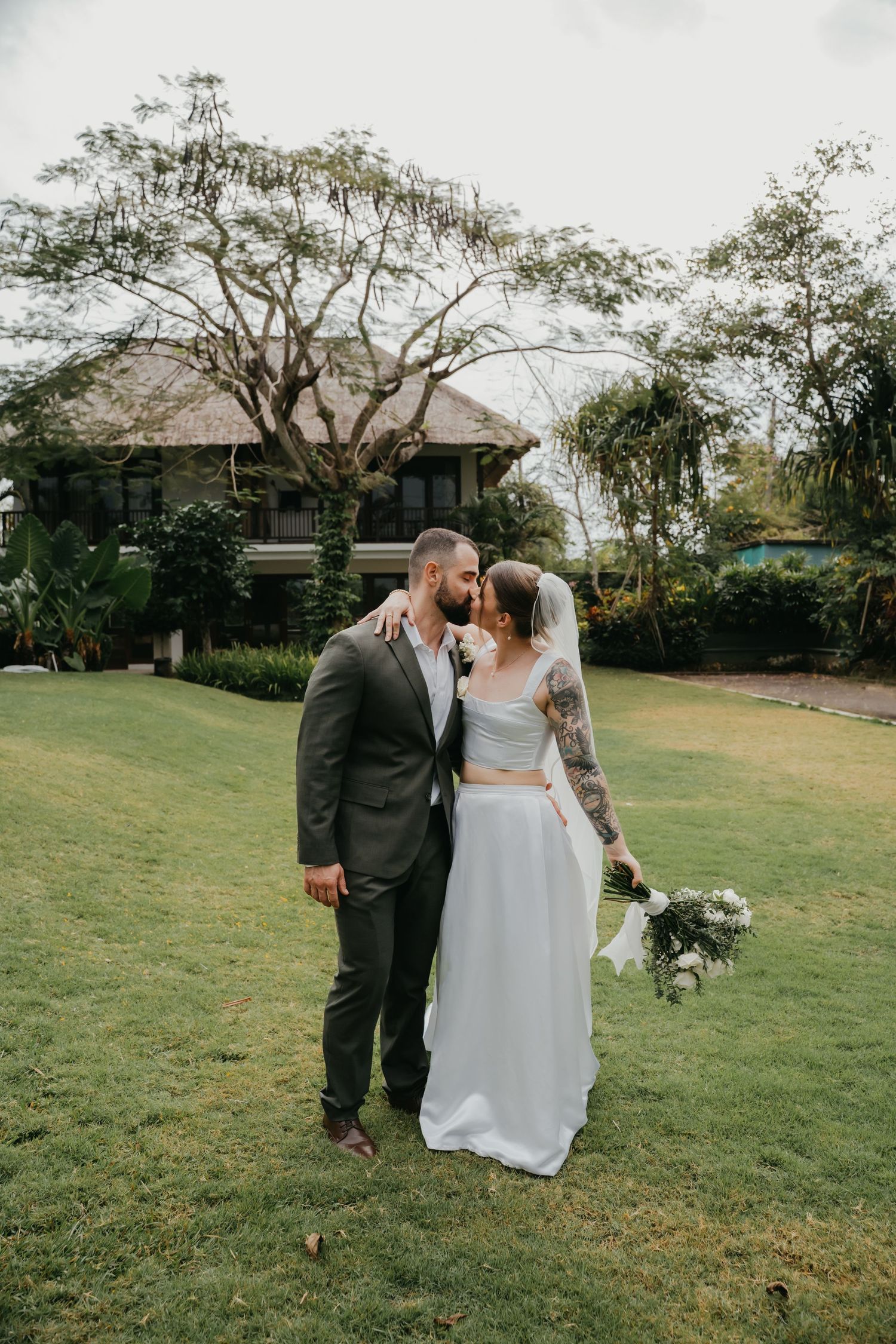 Bride and groom share a kiss on green lawn with tropical trees and villa in background.