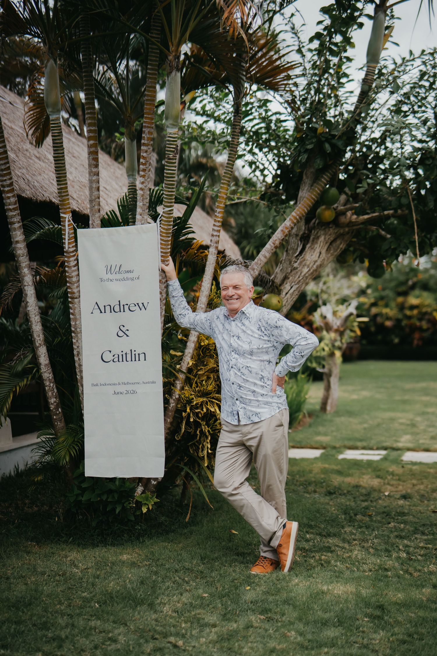 Man in floral shirt posing beside Andrew and Caitlin wedding welcome sign in tropical garden.