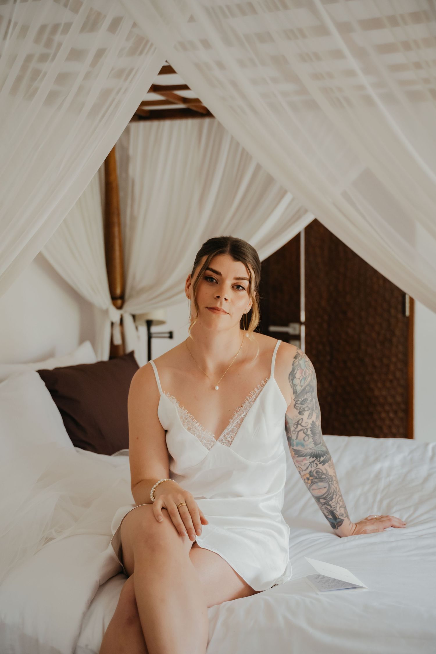 Tattooed bride in white lace slip dress sits serenely on a canopy bed in a tropical villa bedroom.