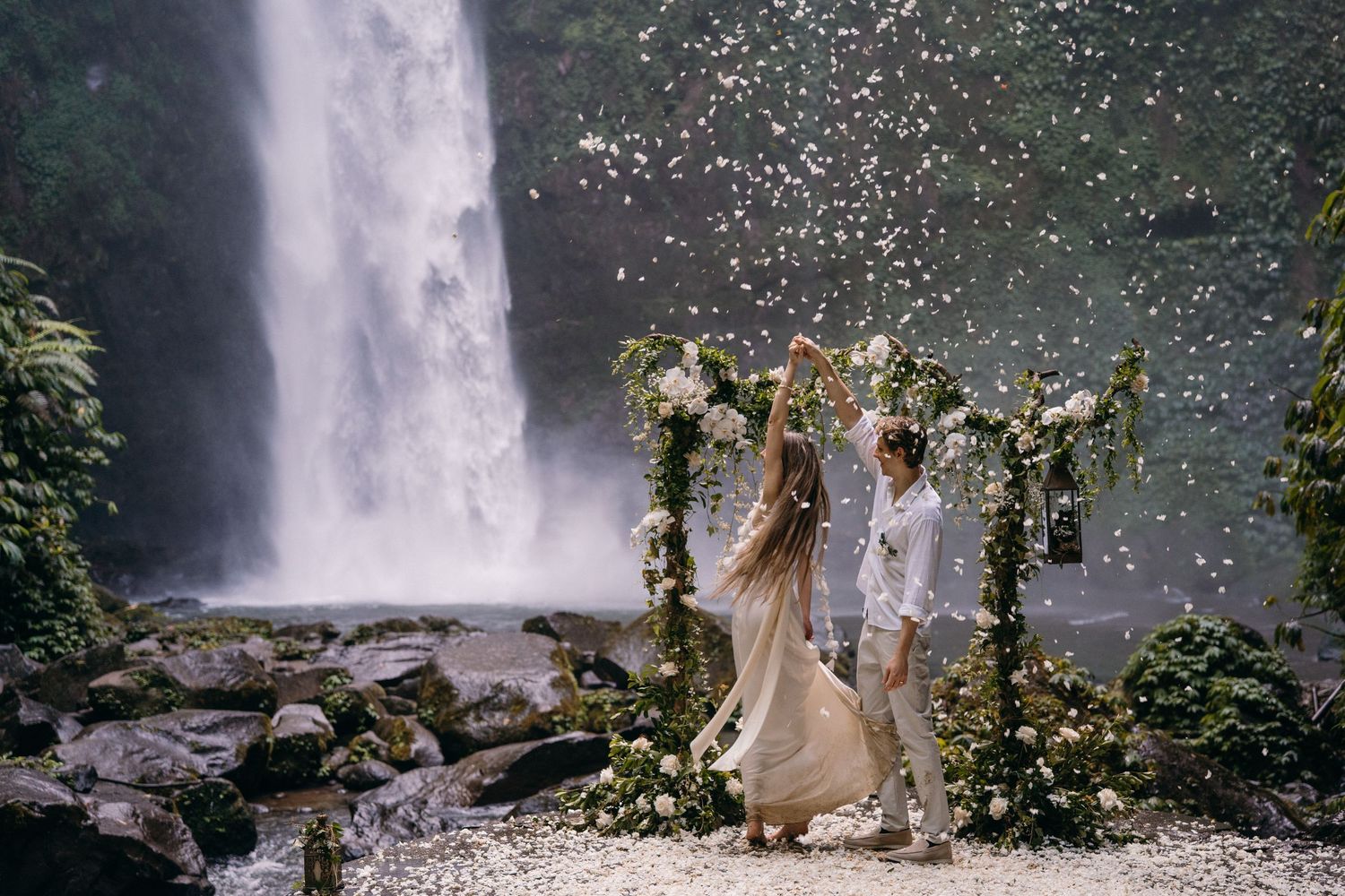 A romantic wedding couple poses in front of a majestic waterfall surrounded by lush greenery and mist.