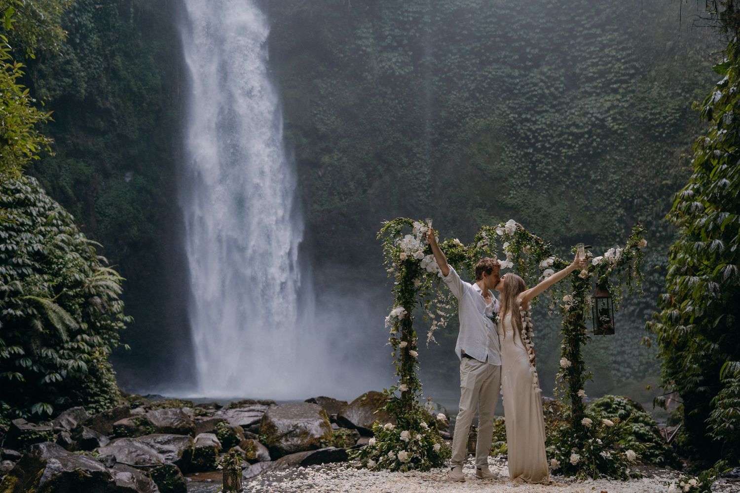 Scenic waterfall cascades down lush green cliffs in a tropical forest setting while people enjoy the view.