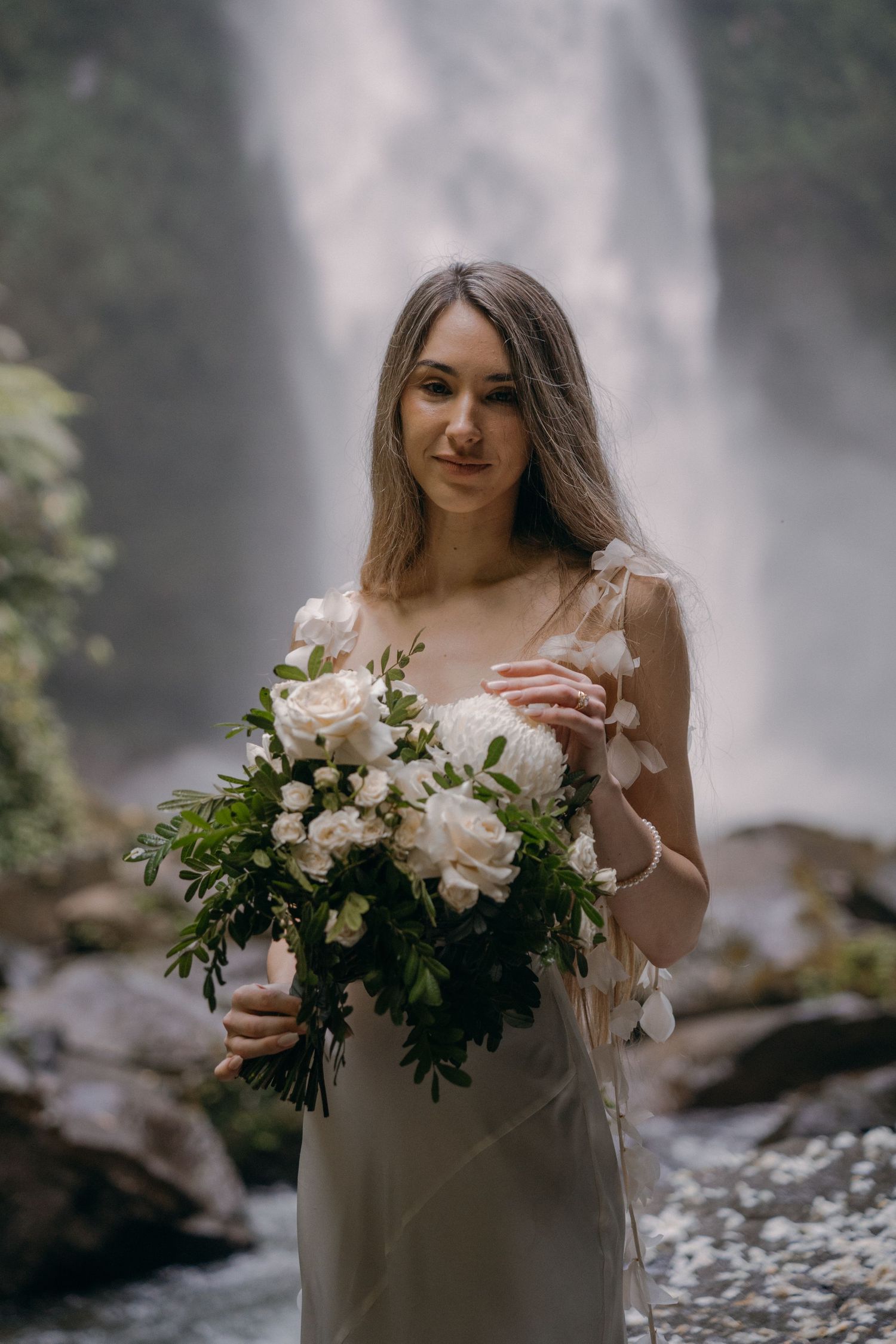 A misty waterfall scene with a person in a light dress holding elegant floral bouquets of white and pink roses.