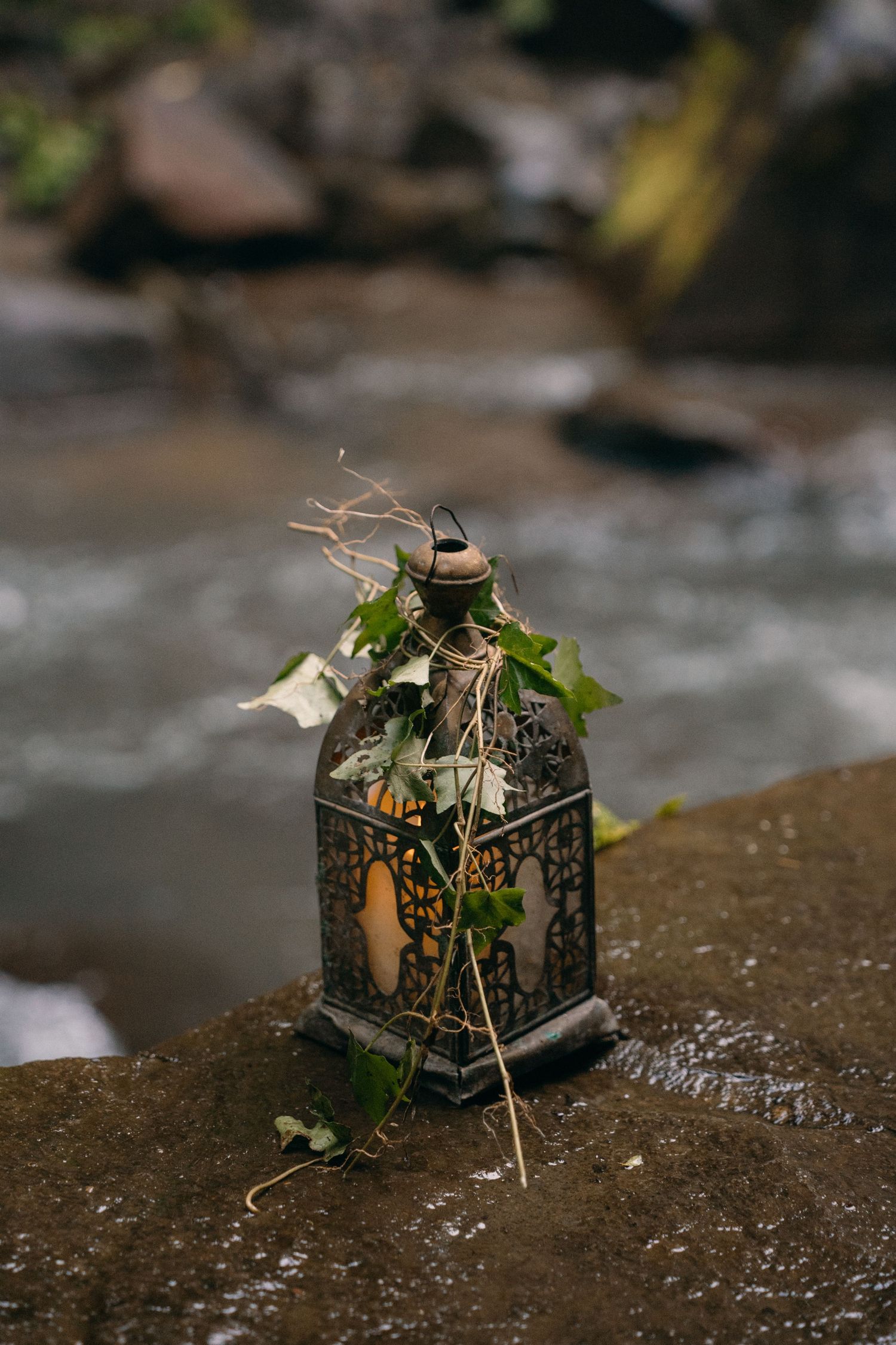 A decorative vintage lantern adorned with leaves and twigs sits on a stone surface near flowing water.