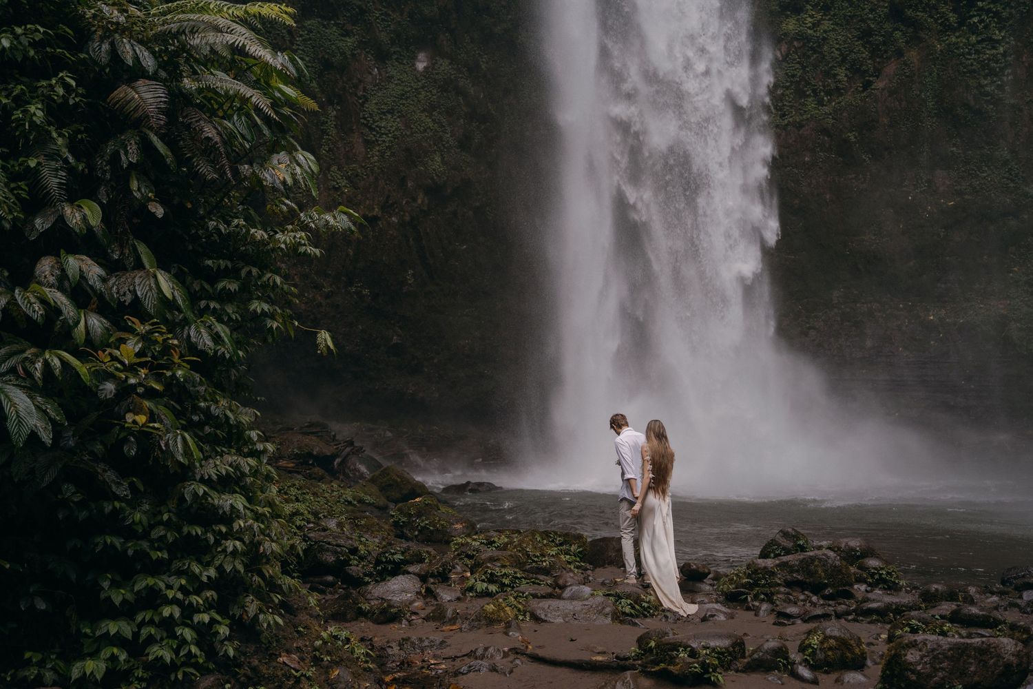 A couple poses together near a majestic tropical waterfall surrounded by lush greenery in a romantic setting.