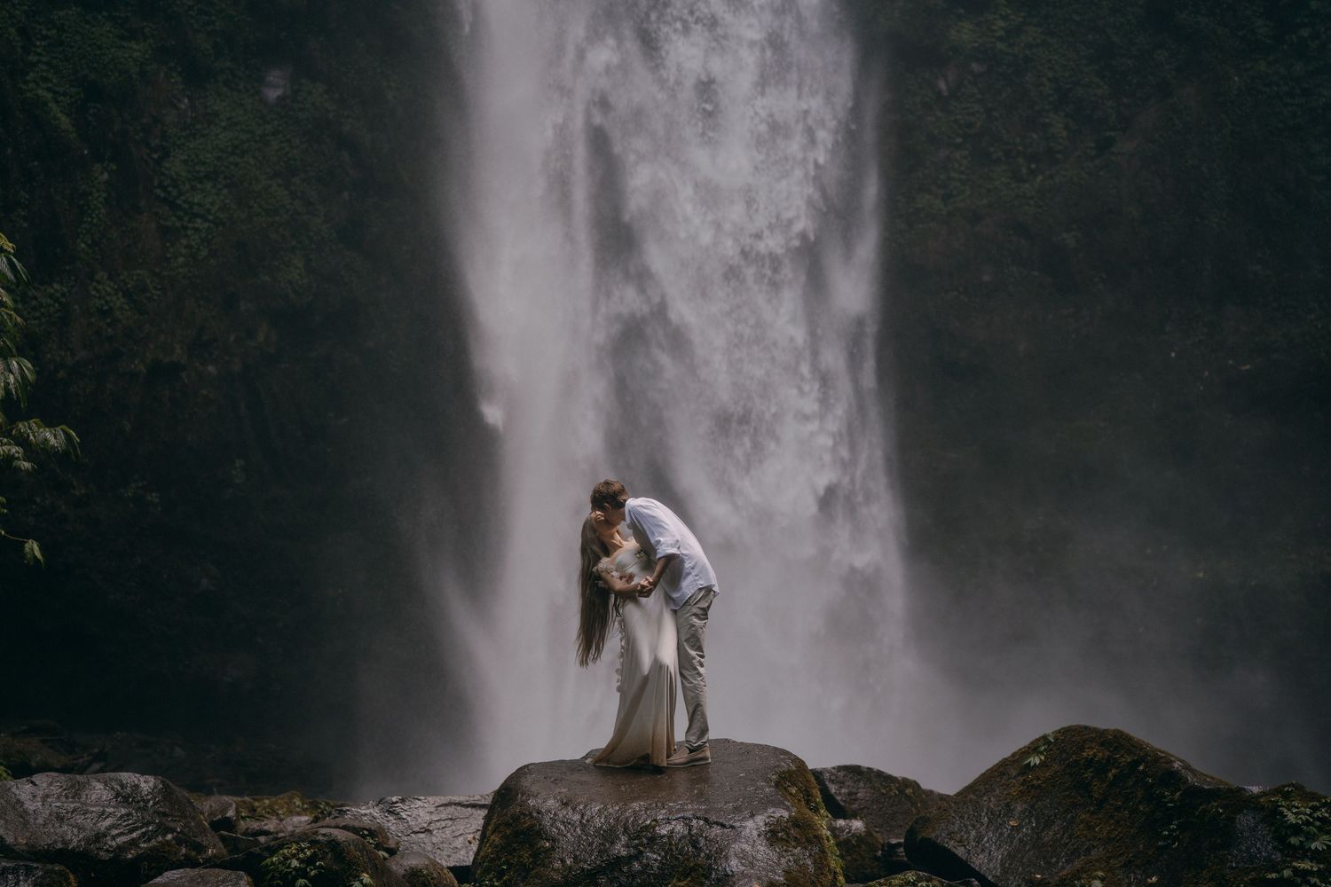 A couple embraces on a rocky outcrop in front of a misty cascading waterfall.