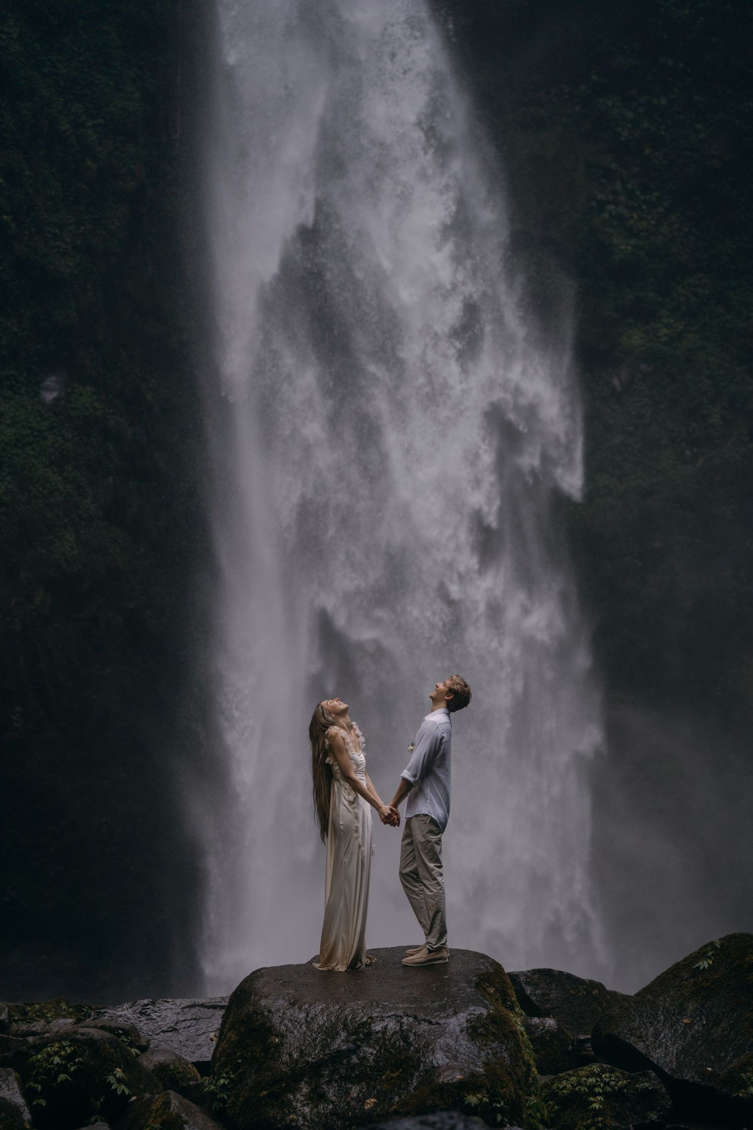 Two silhouetted figures stand against the dramatic backdrop of a towering waterfall in a misty, atmospheric scene.