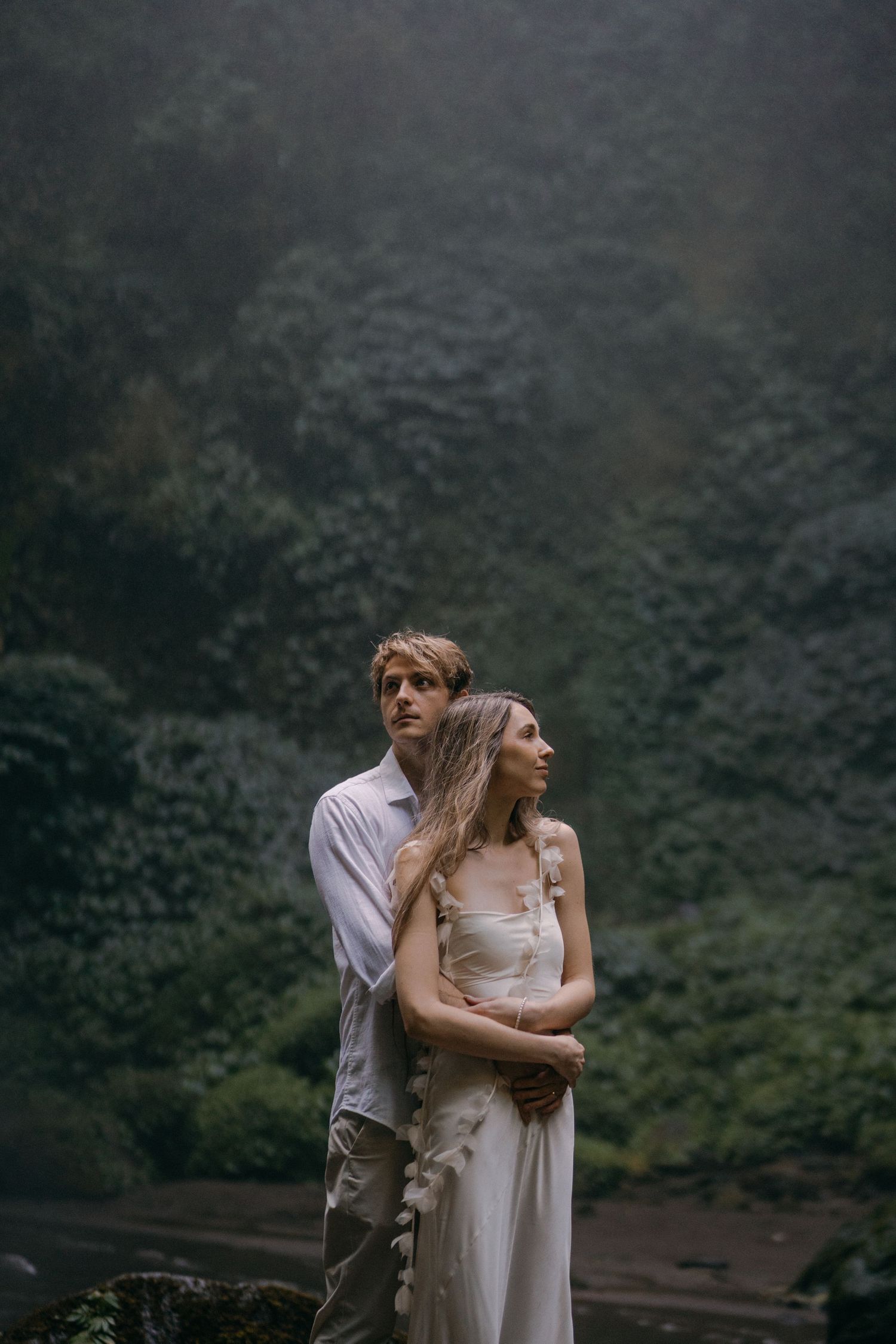 A couple in light colored clothing embrace against a misty mountain backdrop in a romantic outdoor setting.